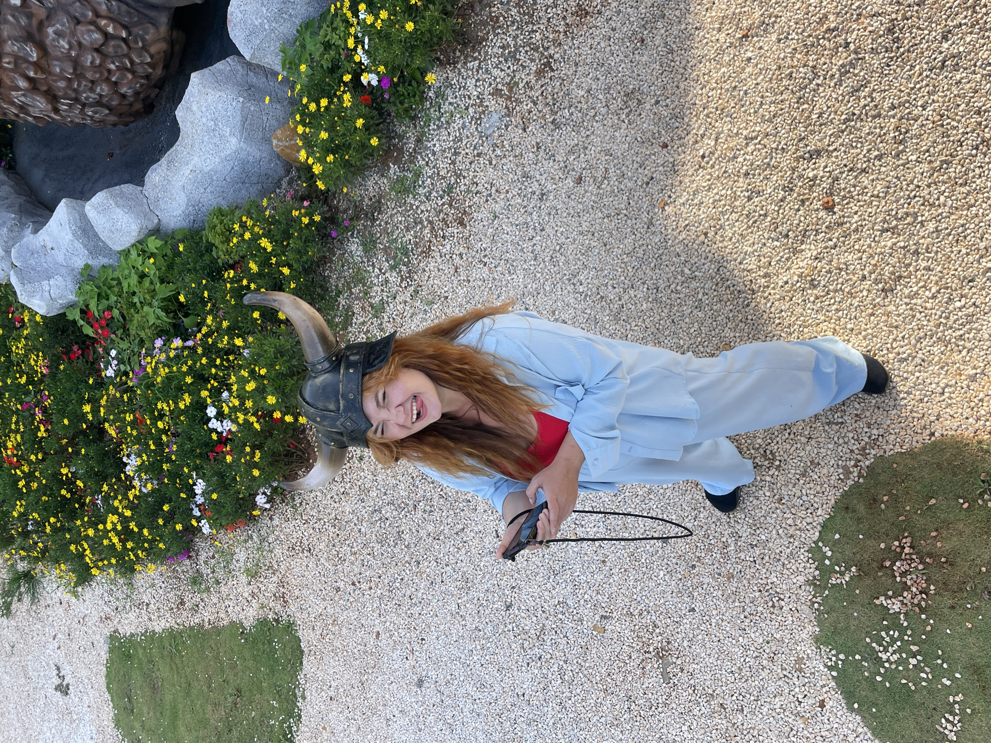 In Thôn 1, Vietnam, a person with long, reddish-brown hair wears a Viking helmet with horns, a red top, and a light blue pantsuit. They are standing on a ground covered with small, light-colored pebbles. The person is holding a black electronic device in their hands and has their tongue sticking out, appearing to be making a humorous expression. To the upper left, a patch of green grass is visible, also scattered with pebbles. In the background, a dense flower bed with numerous yellow, white, and red blossoms is situated next to large, gray, sculpted rocks that form a decorative border.