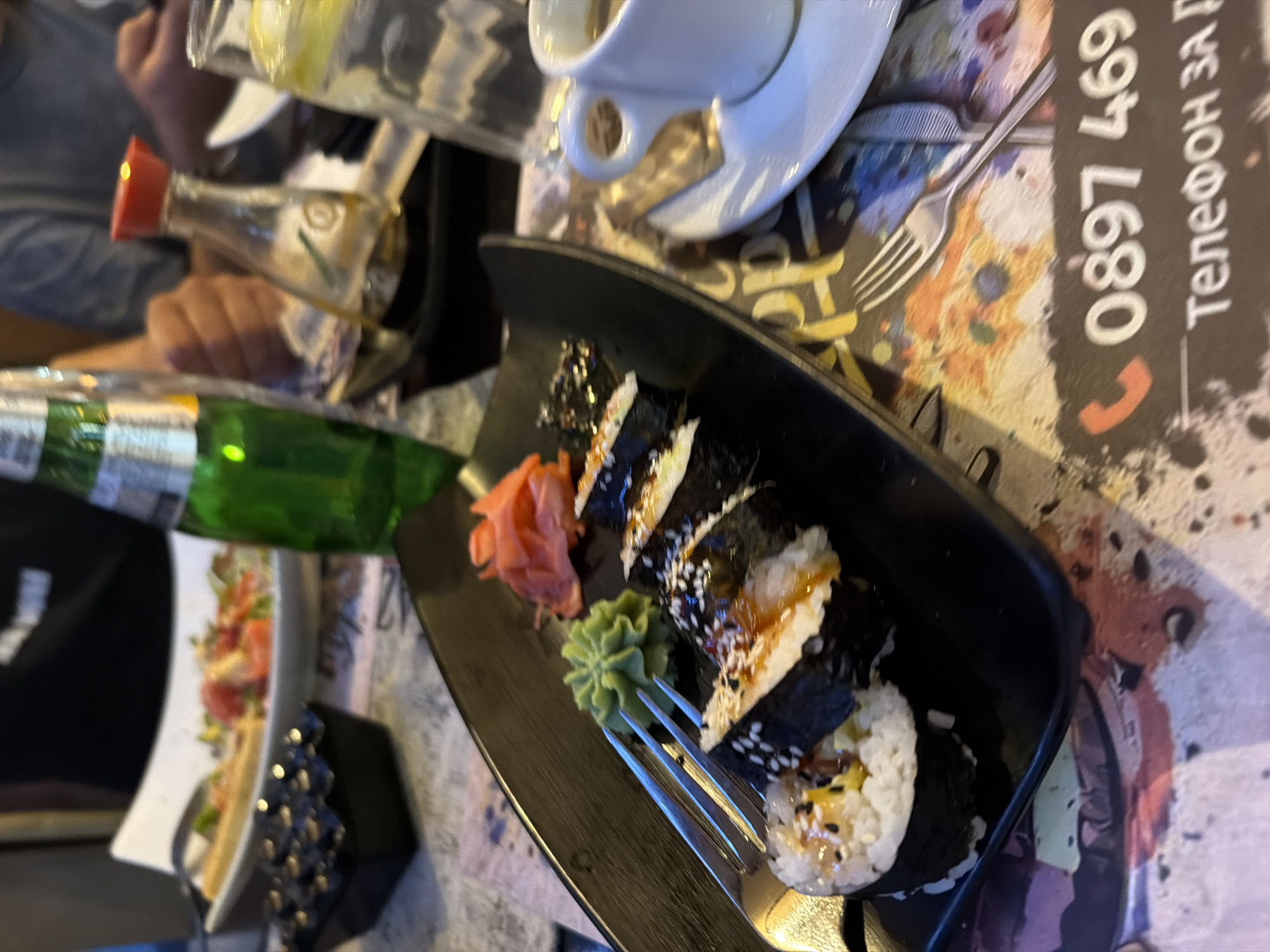 The image presents an overhead, angled view of a dining table, likely in a restaurant or cafe setting. The central subject is a black rectangular plate holding several pieces of sushi rolls (maki) adorned with sesame seeds and a glossy sauce. Accompanying the sushi are a mound of bright pink pickled ginger and a dollop of green wasabi paste. A silver fork rests on the edge of the plate.
Various other objects are scattered across the table. A white ceramic coffee cup on a saucer is visible towards the top right. To the left, a green glass bottle, possibly for a beverage, and a small glass bottle with a red cap, likely soy sauce, are present. A clear glass bottle, presumably water, is also seen. A partially obscured white plate in the lower left background appears to hold a salad or another dish. Two partially visible sets of hands and arms suggest individuals are seated around the table, engaged in a meal.
The table surface beneath the dishes features a colorful, abstract pattern with splashes of brown, blue, yellow, and white. A prominent detail is a black card or menu displaying white text, including the numbers "0897 469" and the Cyrillic phrase "Телефон за" (Telefon za), which translates from Bulgarian to "Phone for". This indicates the location is likely in Bulgaria or a Bulgarian-speaking region. The scene depicts a casual dining experience, with no specific time of day discernible from the indoor lighting.