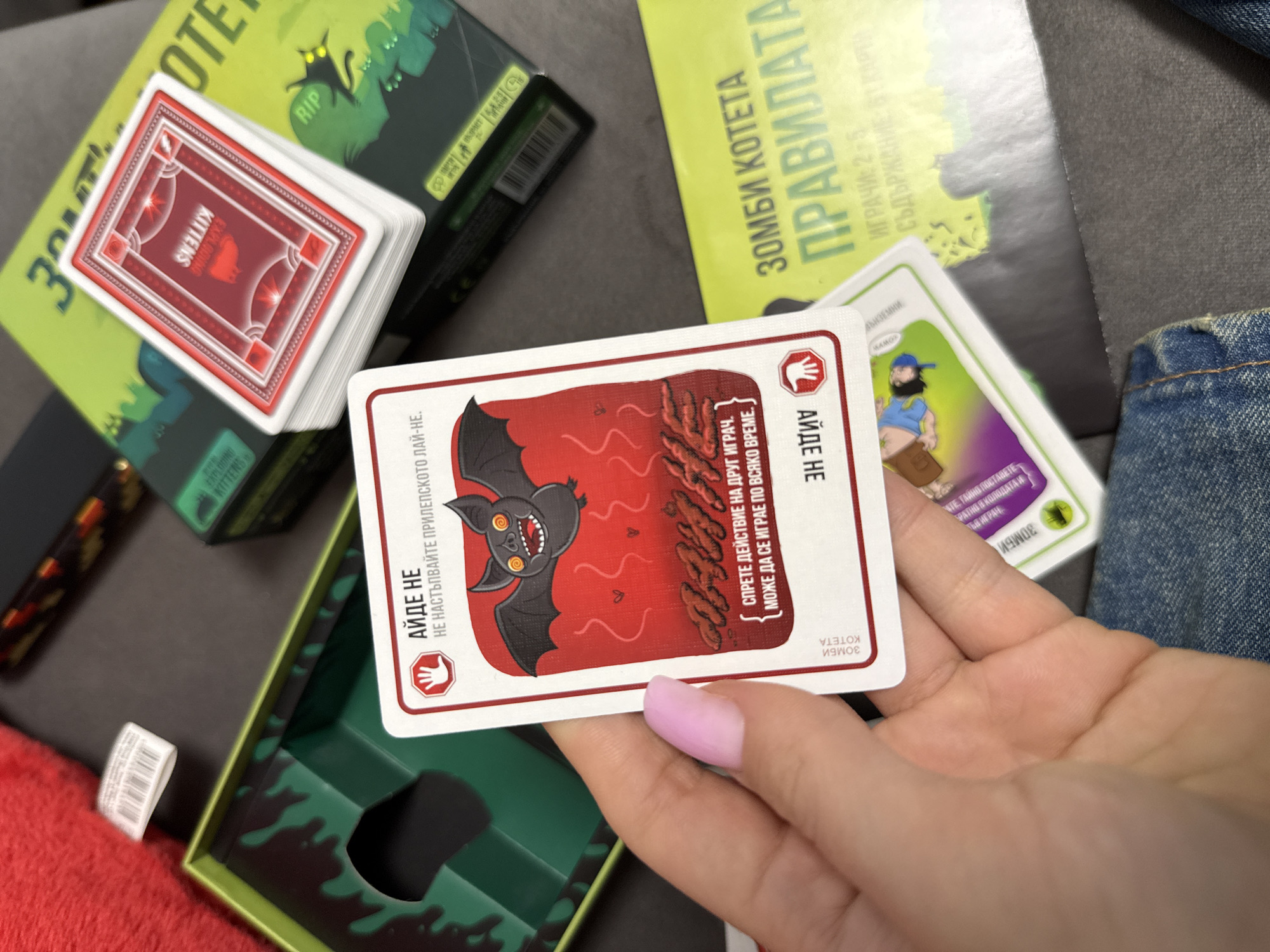 The image shows a person holding a playing card from the game "Zombie Kittens." The card features a cartoon bat and text in Bulgarian. A stack of "Exploding Kittens" cards and game boxes for both "Zombie Kittens" and "Exploding Kittens" are also visible. The scene appears to be indoors on a gray surface, possibly in Sofia, Bulgaria based on the language on the packaging. The person's hand is manicured with a light pink nail polish. A small part of a denim garment is also visible.