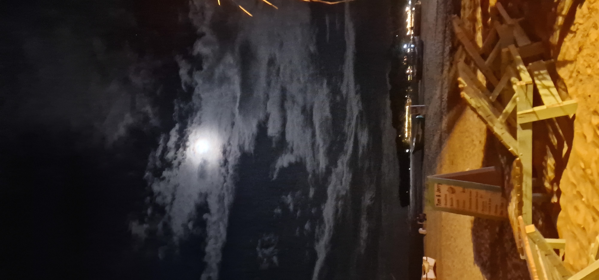 The image presents a serene night scene, likely along the coast of Arugam Bay, Sri Lanka. The sky, occupying the majority of the frame, is dark but illuminated by a bright, prominent moon partially obscured by scattered clouds. The moonlight creates a luminous effect on the clouds, highlighting their shapes and textures against the dark backdrop.
Below the sky, a body of water, presumably the ocean, stretches across the midground, reflecting faint artificial lights from the distant shore. These reflections suggest the presence of a town or establishments further along the coastline.
In the foreground, on the right side of the image, there is a sandy or earthen area bathed in warm, artificial light. Several wooden structures are visible here, appearing to be rustic fences, barriers, or possibly part of a construction or seating arrangement. Near these wooden structures, a stand-up sign or menu board is prominently displayed. The top of the sign clearly reads "Tom & Jerry," suggesting a café, restaurant, or bar. Below this title, a list of items is visible, likely a menu, though the specific text is unreadable due to the distance and image resolution.
The overall impression is one of a peaceful, late-night setting by the sea, characterized by the natural beauty of the moonlit sky and the subtle presence of human activity indicated by the artificial lights and the "Tom & Jerry" establishment. No people are visible in the scene.