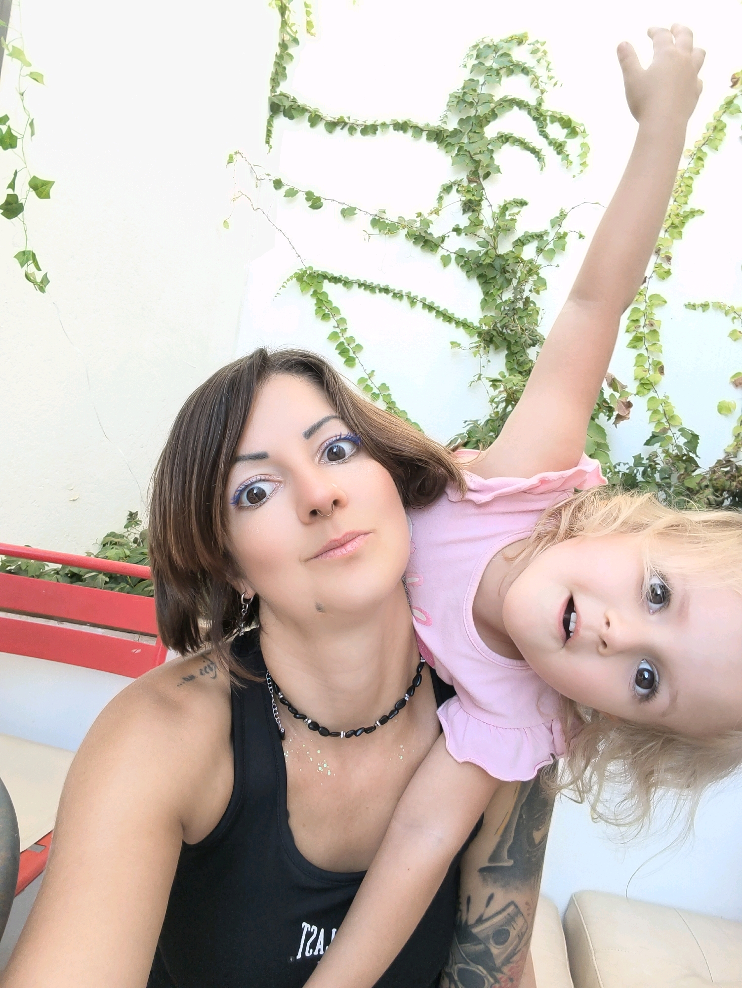 In an outdoor setting, likely within the Lachish Regional Council of Israel, a woman with short brown hair and blue eyeliner is taking a selfie. She is wearing a black tank top and a beaded necklace, and her left arm is tattooed. Her daughter, a young blonde girl in a pink t-shirt, is held upside down on her back. The daughter is reaching her arm up, and both are looking at the camera. Behind them, a white wall is covered in climbing green ivy. A red bench is visible to the left of the frame.