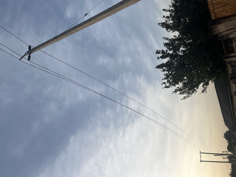 The image captures an outdoor scene in Barnawa, Nigeria, featuring prominent utility infrastructure against a partially cloudy sky. A weathered concrete utility pole, equipped with insulators and multiple dark power lines, dominates the upper right quadrant, extending diagonally towards the lower right. Three main power lines stretch horizontally across the middle of the frame, converging towards the top of the pole. In the lower middle and right, the dark green canopy of a large tree is visible. Below the tree, a dark-colored roofline of a building is partially visible, fronted by a low concrete wall. A rusty, metallic gate is present on the far right, partially obscured by the tree foliage and the wall. In the distant lower left, a second, smaller utility pole with additional power lines is visible against a lighter, hazy sky. The sky transitions from a light blue-grey with scattered white cloud formations at the top to a warmer, lighter hue towards the bottom left, suggesting either early morning or late afternoon conditions. No individuals or specific activities are observable.