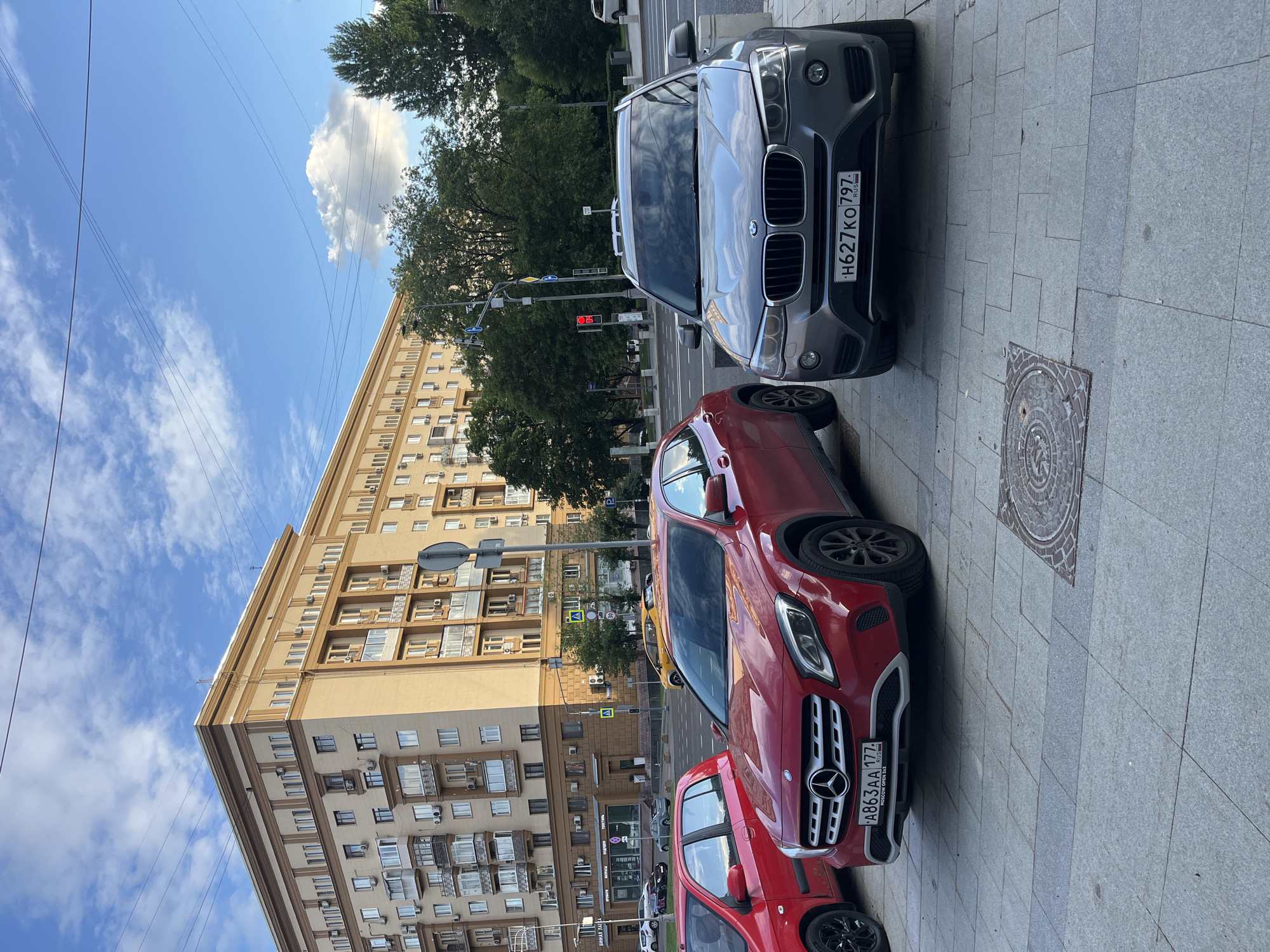 The image depicts a street scene in Moscow, Russia. The primary subjects are several parked cars, including a dark grey BMW SUV and two red Mercedes-Benz vehicles. The BMW has a license plate that reads "H627KO 797". One of the Mercedes-Benz cars has a license plate that reads "A863AA 177". The cars are parked on a sidewalk paved with grey tiles.
In the background, a large, light-colored apartment building with many windows and balconies dominates the scene. Trees line the street in front of the building. Overhead, power lines crisscross the bright blue sky, which is dotted with white clouds. The lighting suggests it is daytime, likely late afternoon or early evening, given the angle of the sun casting shadows. A traffic light showing red is visible, indicating potential traffic flow. The overall impression is of an urban environment on a clear day.