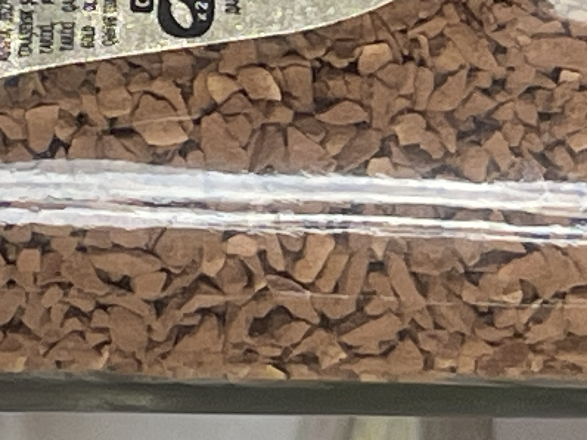 The image is a close-up, vertical shot of instant coffee granules inside a glass jar. The granules are a reddish-brown color and appear to be of irregular, broken shapes, filling most of the frame. A vertical, reflective surface, possibly the glass of the jar itself or a label behind the granules, runs through the center of the image, creating a bright highlight and obscuring some of the coffee. On the right side of the image, partially visible text and graphics on a label can be seen. The text is small and appears to be in a light color against a darker background. The graphics include what looks like a stylized logo with concentric circles and the number "2" or "x2". The overall lighting is soft, with the focus on the texture and color of the coffee. No specific location in Tashkent, Uzbekistan, can be identified from the visual information provided.