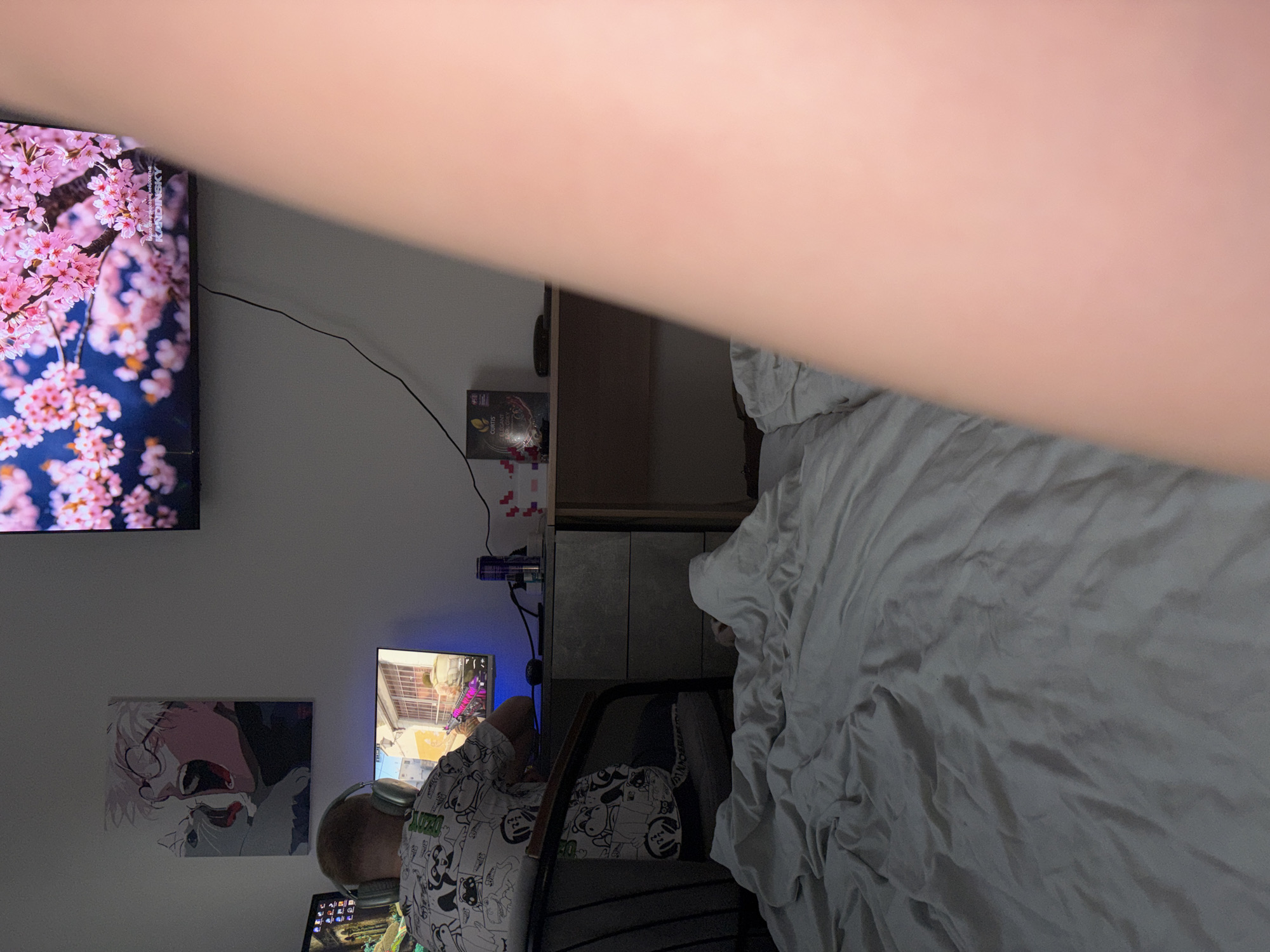The image captures an indoor room, likely a bedroom or personal entertainment space, under dim lighting, suggesting evening or nighttime. A large, blurry, light-skinned arm or body part partially obstructs the top right and center foreground of the shot.
In the mid-ground, a person is seated with their back to the camera, wearing headphones and a white t-shirt patterned with black cartoon figures, including the repeated text "ANZO". They are actively engaged with a computer or gaming setup, as evidenced by the screen in front of them displaying what appears to be a first-person shooter video game.
Two main screens are visible. A large TV mounted on the left wall showcases a vibrant image of pink cherry blossoms (sakura) against a dark blue background. Text on this screen reads "KANDINSKY" and "ТОЛЬКО РАБОТЫ ХУДОЖНИКА". Below this TV and to the left of the seated person, a wall-mounted canvas print or poster features an anime character with white hair and red eyes.
To the right, a bed with rumpled white bedding occupies a significant portion of the frame. Between the person and the bed, a dark wooden shelf unit or cabinet is visible. On a surface near the large TV, a box with "CURVES" and "SMART" printed on it can be seen next to a black cable. Other details include a purple cylindrical object and small colorful square stickers on the wall above the gaming monitor. The primary light sources are the illuminated screens.