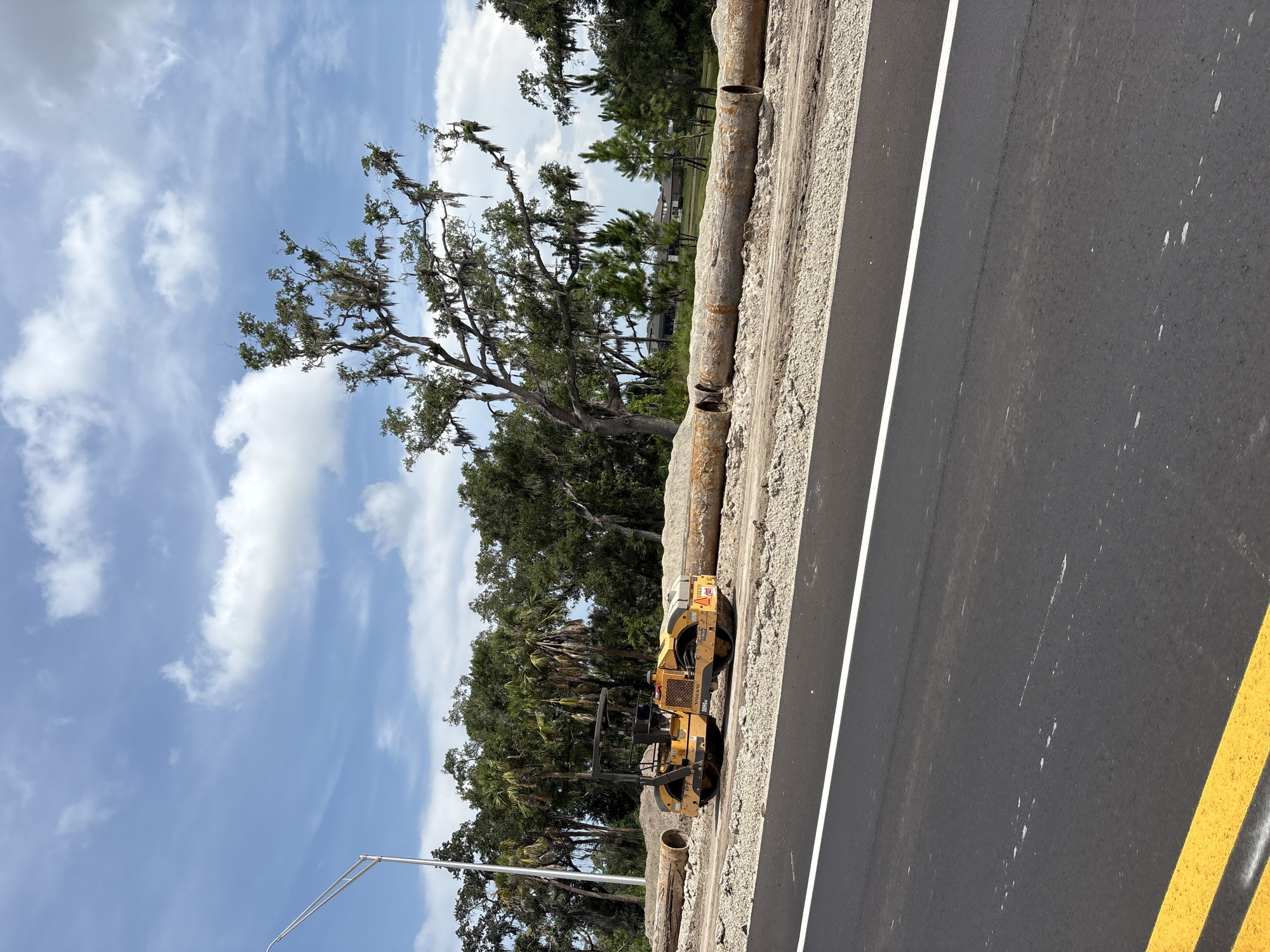 The image shows a roadside construction scene in Parrish, United States. A yellow Volvo roller, a piece of heavy machinery used for compacting surfaces, is parked on the side of an asphalt road that is partially constructed. The road has a white line marking its edge, and a double yellow line is visible on the right side of the image.
The background features lush green trees, including palm trees and mature oak trees draped with Spanish moss, suggesting a warm, humid climate. The sky is a bright blue with scattered white clouds, indicating it is daytime, likely late morning or early afternoon. In the distance, hints of houses can be seen through the trees, suggesting a semi-rural or suburban setting.
The construction activity is evident from the roller, the exposed soil and gravel beside the road, and what appear to be sections of large pipe laid along the embankment. The scene appears peaceful and still, with no people visible. The overall impression is one of ongoing infrastructure development in a natural, sun-drenched environment.