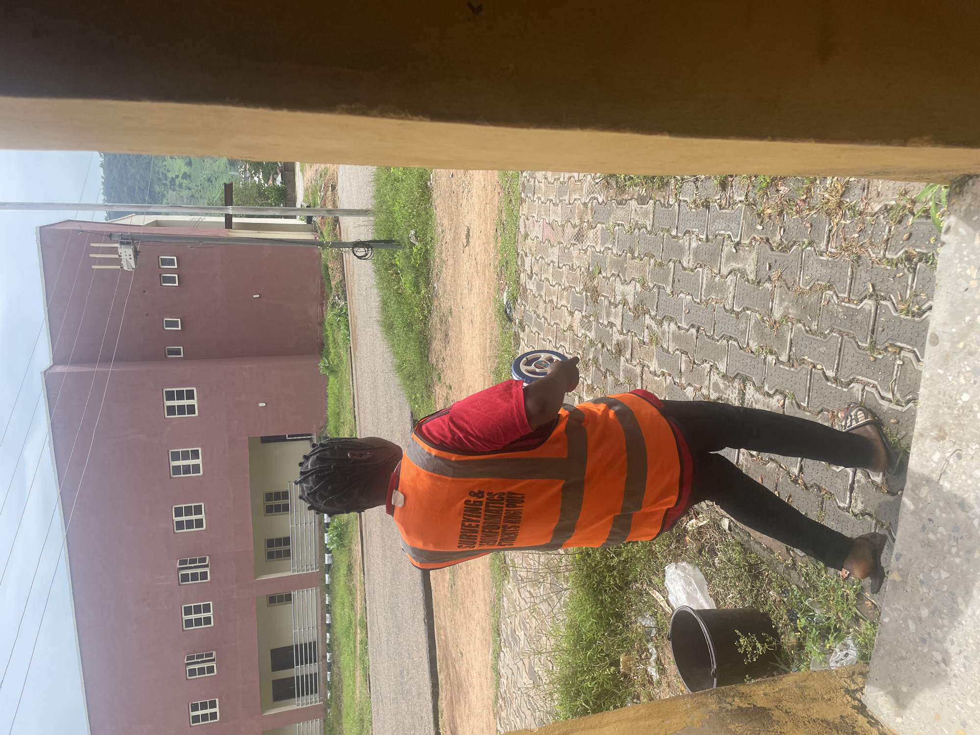 A person wearing an orange high-visibility vest over a red shirt and black leggings stands on a paved walkway in front of a maroon building. The vest has reflective stripes and text that reads "SURVEYING & GEOMATICS STUDENTS KOGI POLY". The person is holding a measuring wheel. To the left of the walkway is a black bucket and some white plastic sheeting. The building appears to be a multi-story structure with many windows. In the background, there is a road, grassy areas, and a hill covered in vegetation. The overall scene suggests an outdoor environment within a campus or urban setting.