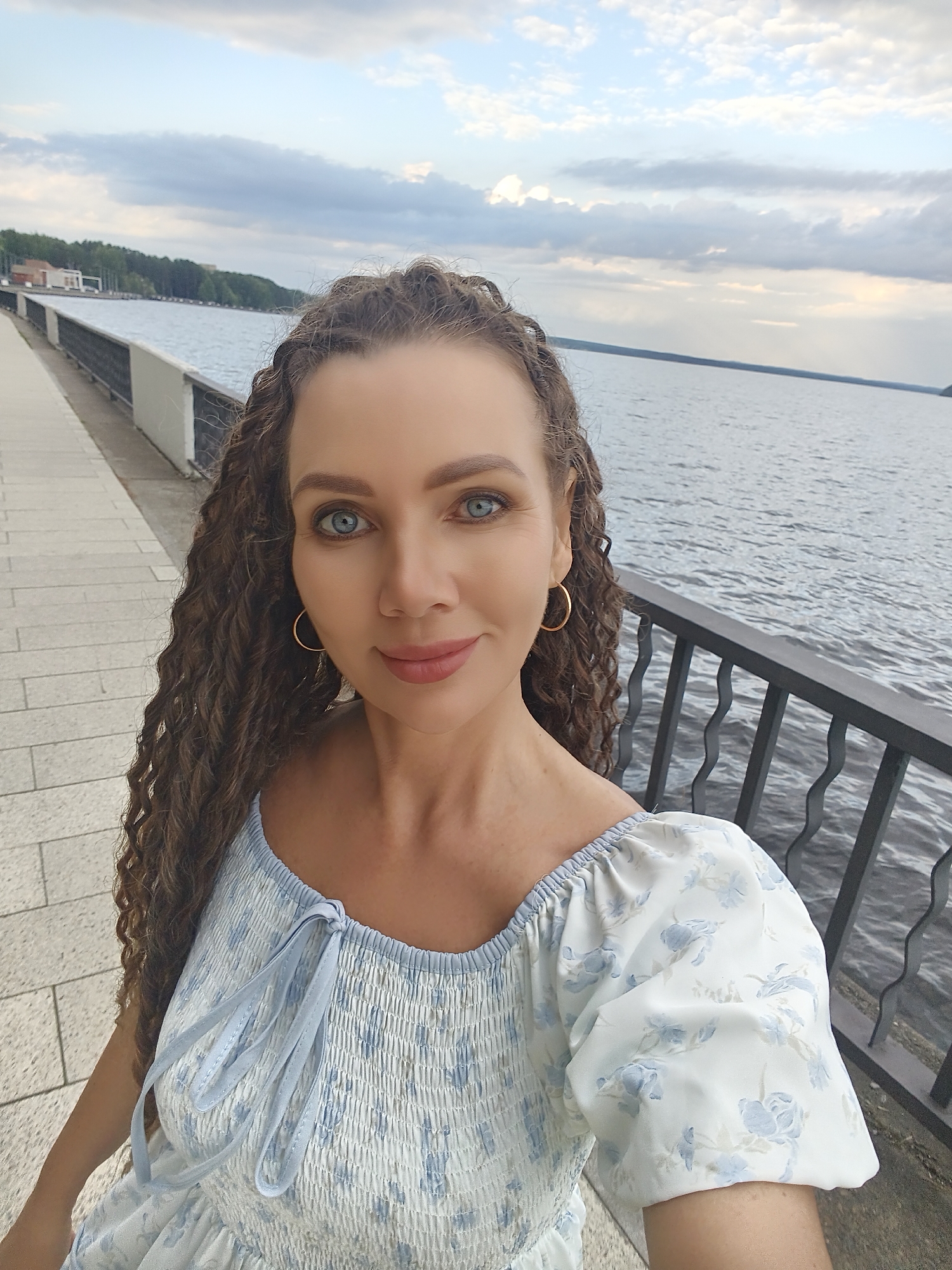 A woman with long, curly brown hair and blue eyes is taking a selfie outdoors next to a body of water. She is wearing a light blue and white floral dress with puffed sleeves. The background shows a paved walkway, a black metal railing, and a wide river or lake with a cloudy sky above. The location is on the embankment in Chaikovsky, Russia.