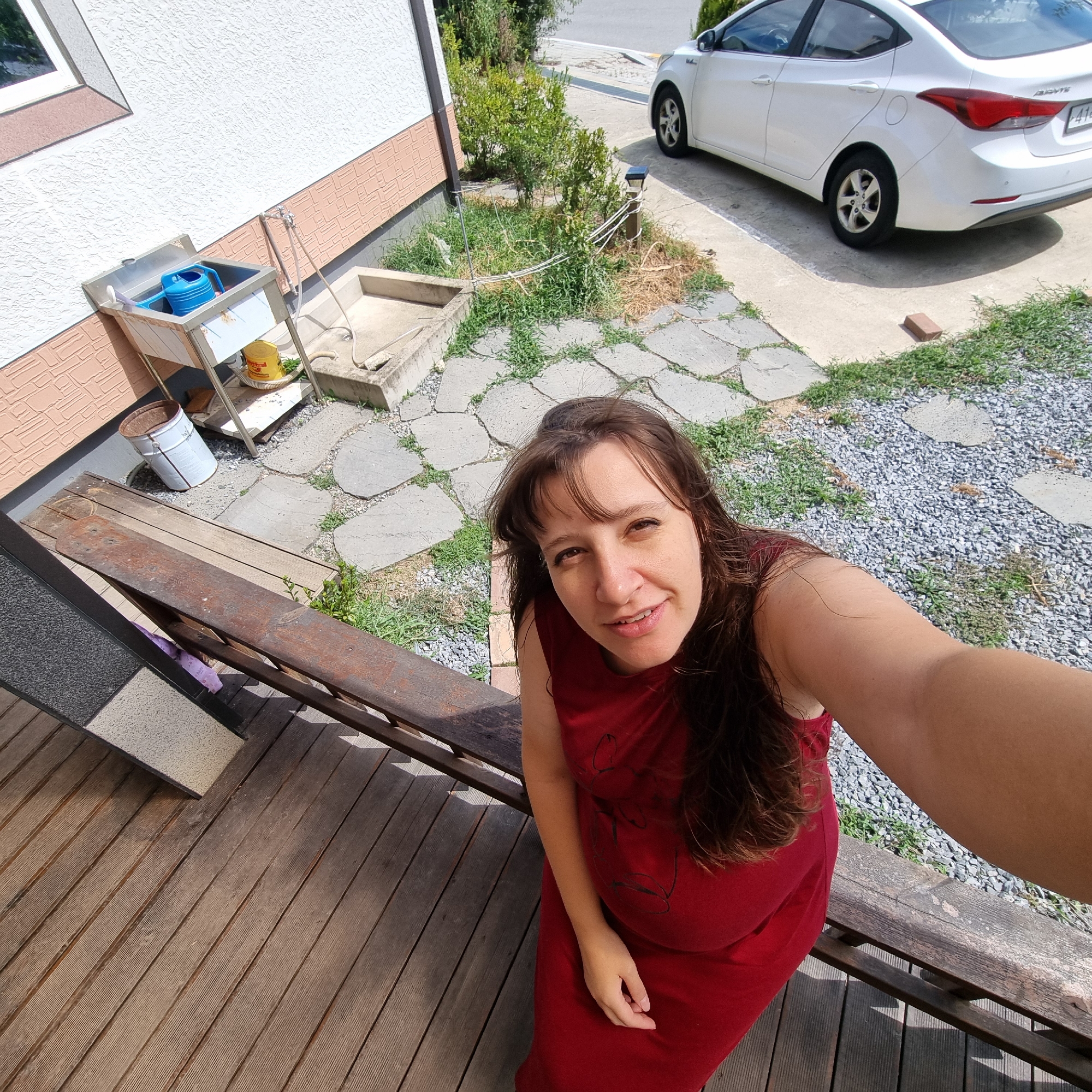 A woman in a red dress is taking a selfie from a wooden deck. The deck has a railing made of weathered wood. In the background, a white car is parked on a concrete driveway. To the left of the driveway, there is a stone sink with a blue watering can. Further left, a white building with textured walls is visible. The ground is covered with gravel and scattered stones. The scene appears to be in a residential area, possibly Dunnae-myeon, South Korea.