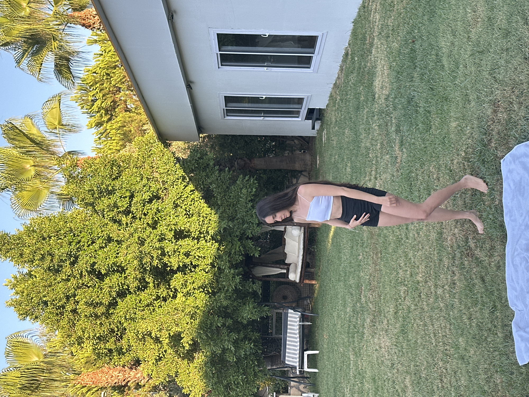 A young woman stands in a grassy backyard in Ortaca, Turkey. She is wearing a white crop top and black shorts, with her hand on her stomach. Behind her, there are palm trees, a large leafy tree, and a white building with two large windows. In the background, there is a bench swing and a daybed. The lighting suggests it is late afternoon.