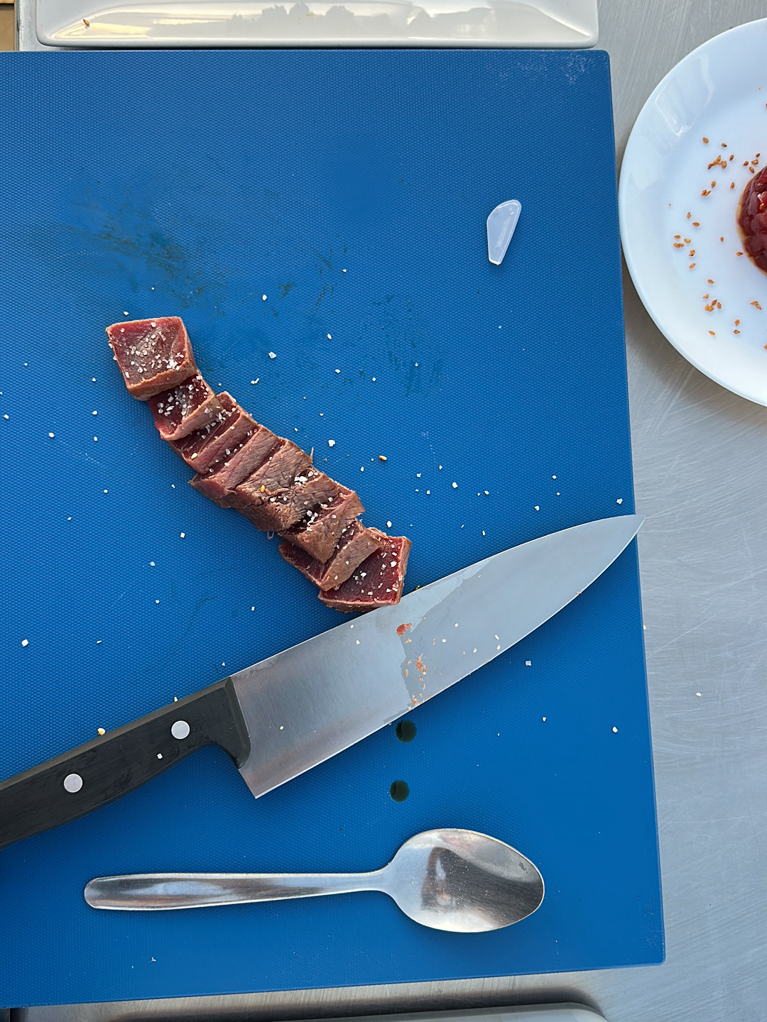 A blue cutting board with sliced meat sprinkled with salt, a knife, and a spoon. A white plate with a mound of meat and salt is partially visible in the upper right corner. The image was taken in Barbate, Spain.