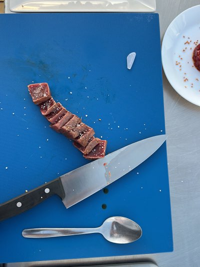 A blue cutting board with sliced meat sprinkled with salt, a knife, and a spoon. A white plate with a mound of meat and salt is partially visible in the upper right corner. The image was taken in Barbate, Spain.