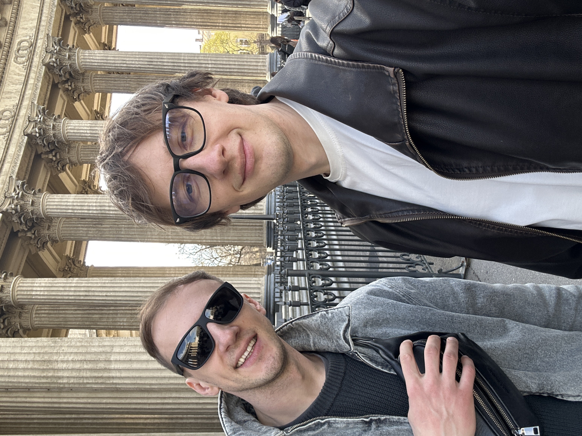Two men pose for a selfie in front of the Kazan Cathedral in Saint Petersburg, Russia. The background features the cathedral's colonnade. The man in the top of the frame wears eyeglasses, a white t-shirt, and a black leather jacket. The man in the bottom of the frame wears sunglasses and a gray denim jacket over a dark shirt. A black bag is partially visible in the lower right corner.