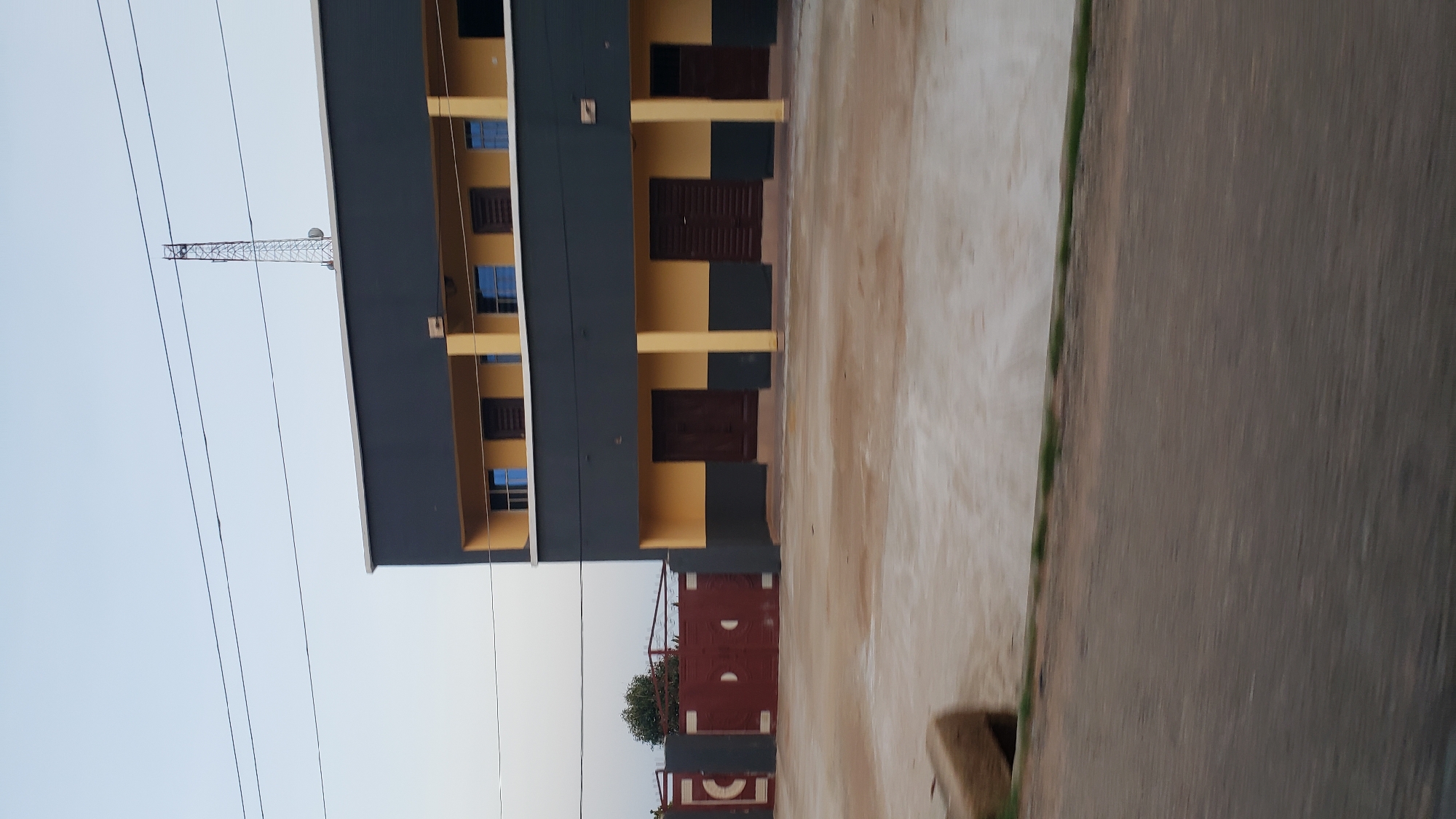 The image depicts a building in Jalingo, Nigeria, with a large, dark gray facade and a lighter yellow lower section. The building has a balcony on the upper level with several windows, and the lower level features multiple dark wooden doors. To the left of the building is a dark red, ornate metal gate, partially obscured by the building. The ground in front of the building appears to be a light-colored, dusty surface, with a dark gray road visible in the foreground, blurred due to motion. A communication tower is visible on the roof of the building in the upper right portion of the image, along with power lines stretching across the sky. The sky is a pale, overcast blue. There are no people or animals visible in the image.