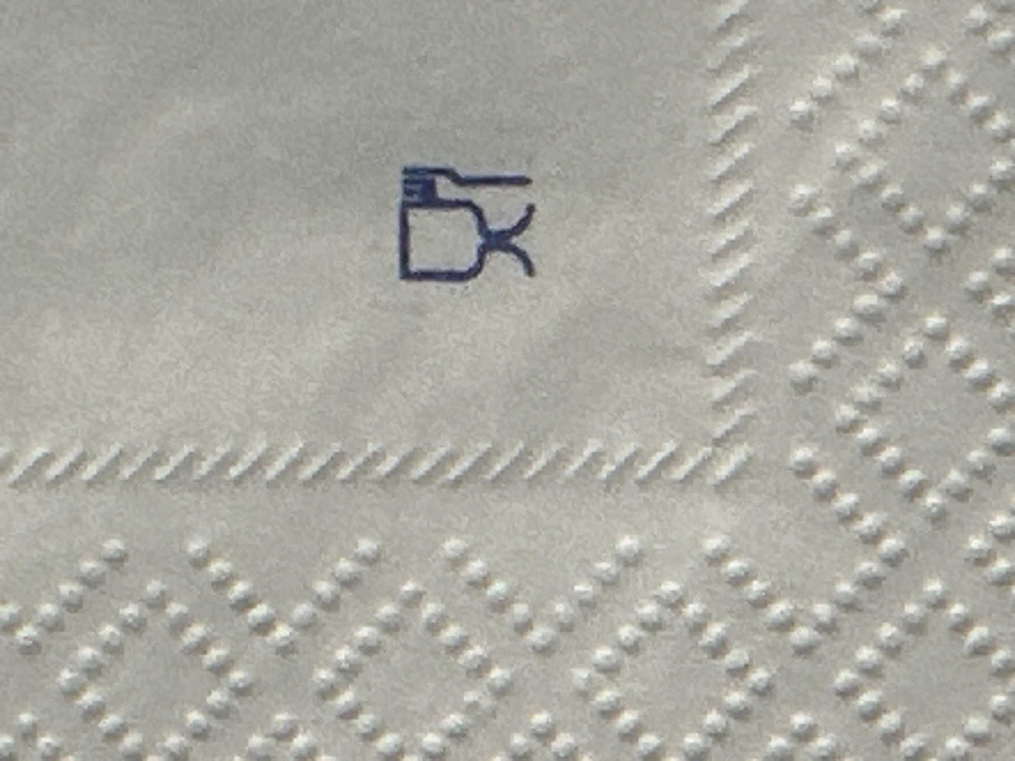 The image presents a close-up, high-magnification view of a textured, light-colored paper or fabric surface, likely a disposable napkin. The material features an embossed or debossed pattern: a grid of small, raised or recessed dots forming diamond shapes across the left side, and a vertical strip of diagonal parallel lines acting as a border along the middle-right. In the upper right quadrant of the surface, a dark blue graphic symbol is printed, depicting a stylized goblet or wine glass to the left of a stylized four-tine fork. This symbol typically indicates food-grade safety or suitability for contact with foodstuffs. No people, discernible actions, or broader environmental context are visible. The specific location within Milan, Italy, or any other city, cannot be determined from the visual information provided in the image.