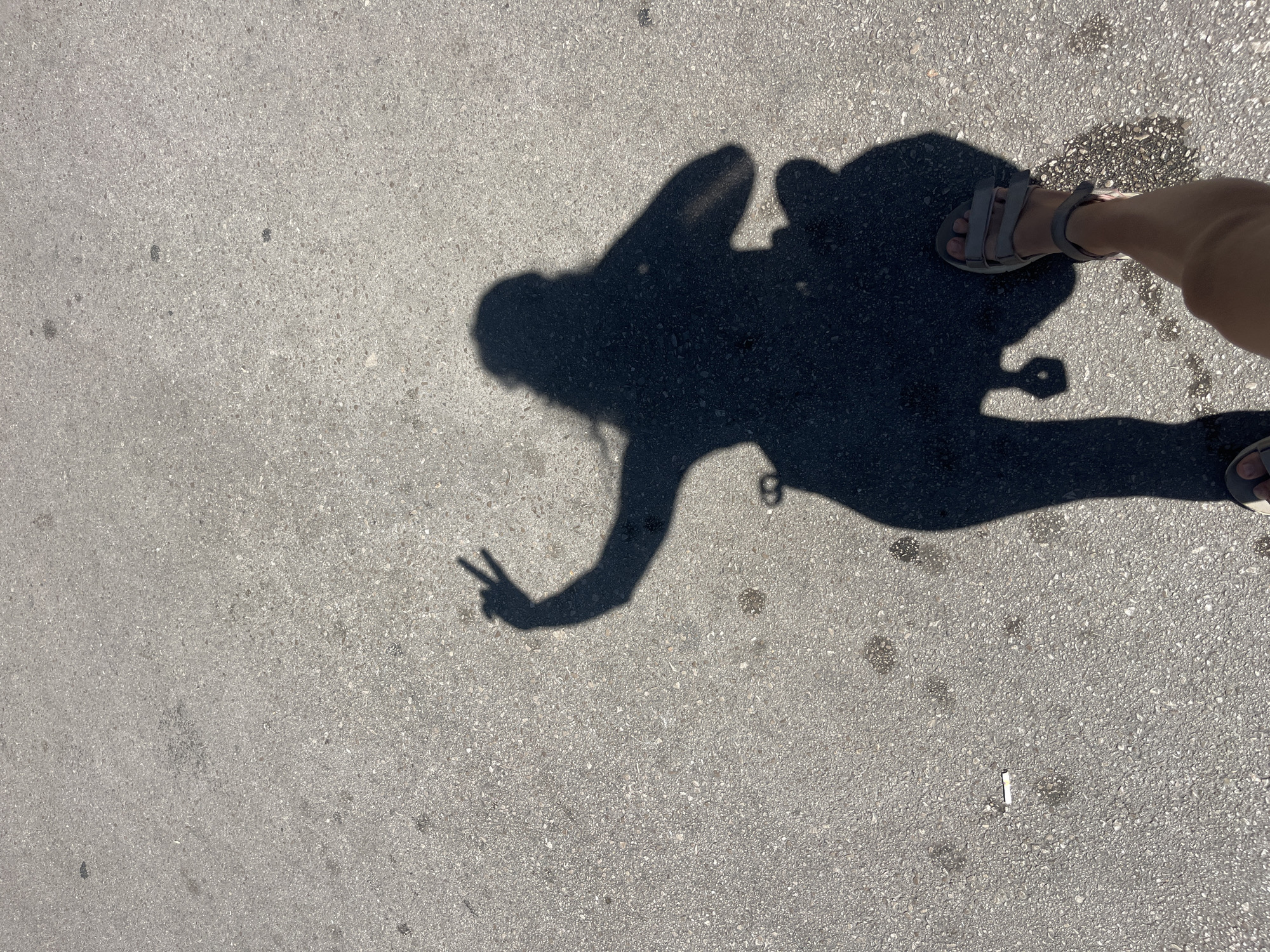 A shadow of a person is cast on an asphalt surface. The shadow is that of a person making a peace sign with their right hand. The person is wearing sandals. There are some dark spots and a cigarette butt on the ground.