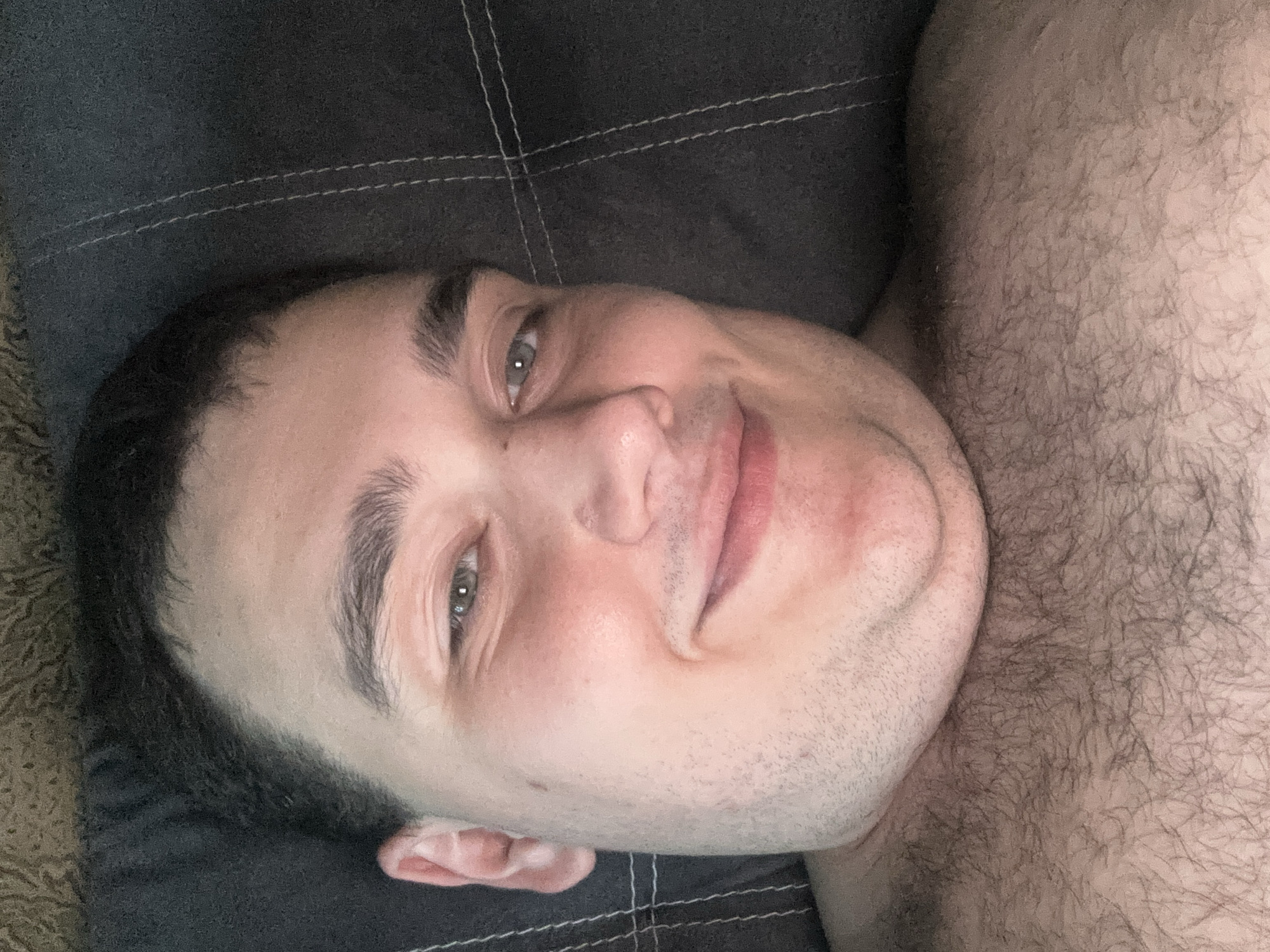 The image is a close-up, overhead shot of a person's face and upper chest. The person appears to be a young adult male with fair skin. He has dark hair that is short on the sides and longer on top, with some hair visible on his chest and shoulder. His eyes are open and he is looking towards the camera with a slight smile. He has stubble on his chin and jawline. The background is dark and appears to be a textured surface, possibly a couch or bed, with visible stitching lines. The lighting is soft and diffused, suggesting it may be indoors, possibly in the evening or on an overcast day. No text is visible in the image.
