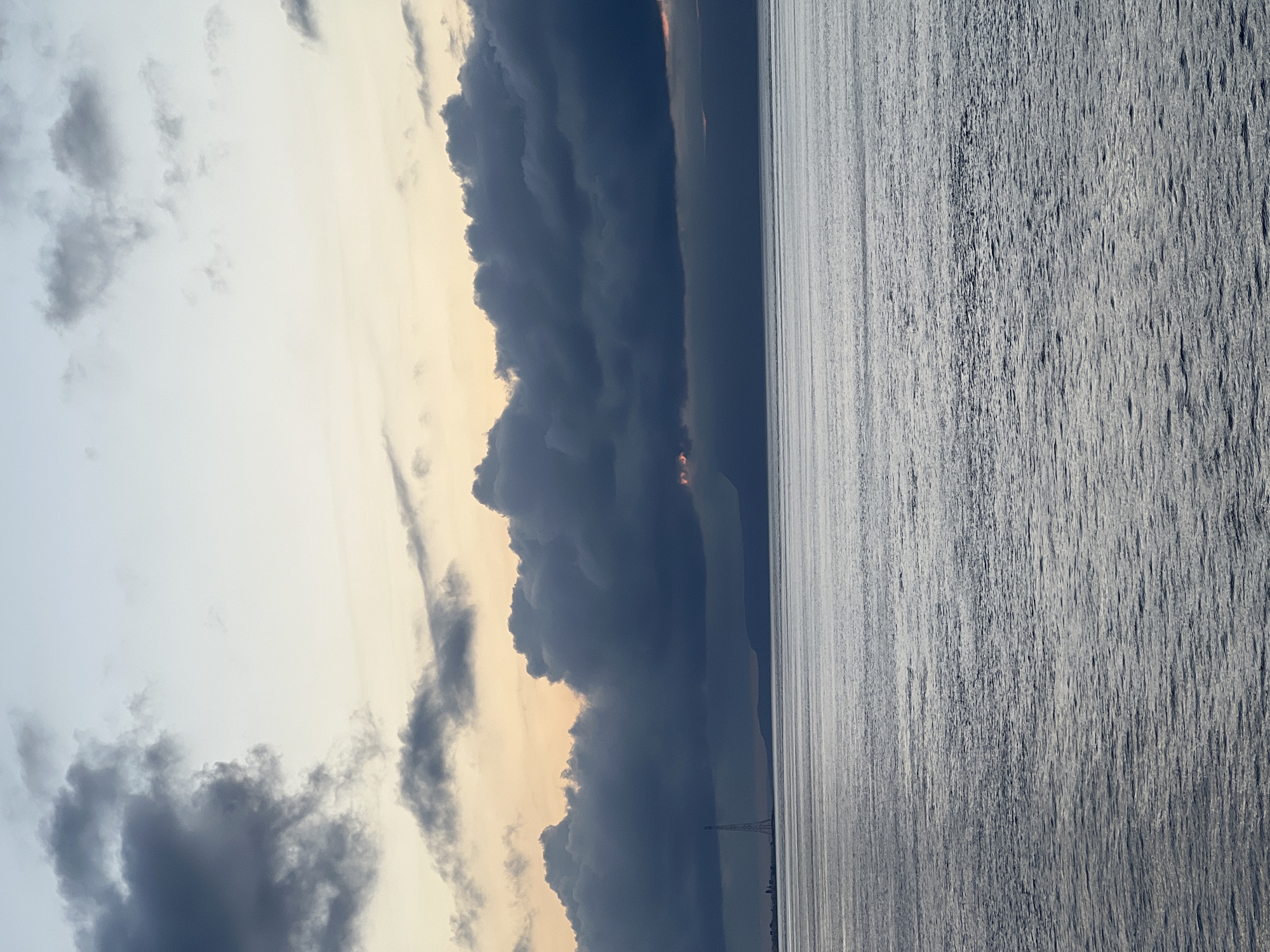 The image presents a panoramic view across the Strait of Messina at either sunrise or sunset. The sky in the upper third of the frame transitions from a light blue-grey at the zenith to pale yellow and soft orange hues near the horizon. A broad, dense band of dark grey to almost black cumulonimbus clouds dominates the middle ground, extending horizontally across the sky and obscuring portions of the distant land. Pockets of vibrant orange and pink light are visible breaking through the gaps in these dark clouds, particularly on the right side and near the center-left.
The lower two-thirds of the image depicts the surface of the Strait of Messina, rendered in shades of shimmering silvery-grey. The water is textured with numerous small ripples and waves, creating a subtle, reflective pattern across its expanse, indicating calm but dynamic conditions.
Along the distant horizon, beneath the heavy cloud band, a dark silhouette of land, likely the coast of Calabria, is visible. On the left side of this distant shoreline, identifiable as part of Messina, a very tall, slender lattice structure—the Pylon of Messina (Torre Faro)—stands prominently. A small cluster of low-rise, indistinct buildings is situated on the distant coast to the immediate left of this tower. No individuals or specific actions are discernible within the scene. The overall lighting suggests a subdued and dramatic atmosphere.