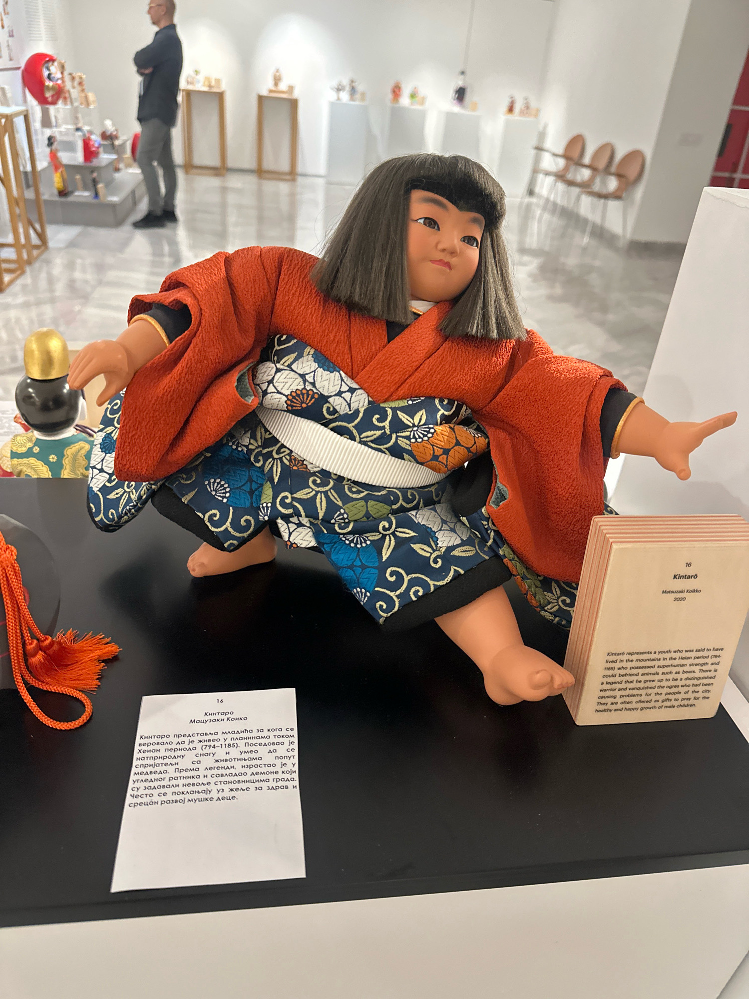 This image shows a traditional Japanese Kintaro doll displayed on a black platform in what appears to be an exhibition space. The doll is dressed in an orange kimono over a blue and white patterned garment, with its legs bent and arms outstretched. To the right of the doll is a small, wooden information card with text in both English and Cyrillic script. The English text describes Kintaro as a youth with superhuman strength who befriended animals and defeated ogres, and mentions that such dolls are offered as gifts to pray for the healthy growth of male children. In the background, out of focus, are various other display items, white walls, and a man standing with his arms crossed, observing the exhibit. The overall environment suggests an indoor art gallery or museum. The specific location in the city of Belgrade, Serbia, cannot be determined from the visual information provided in the image.