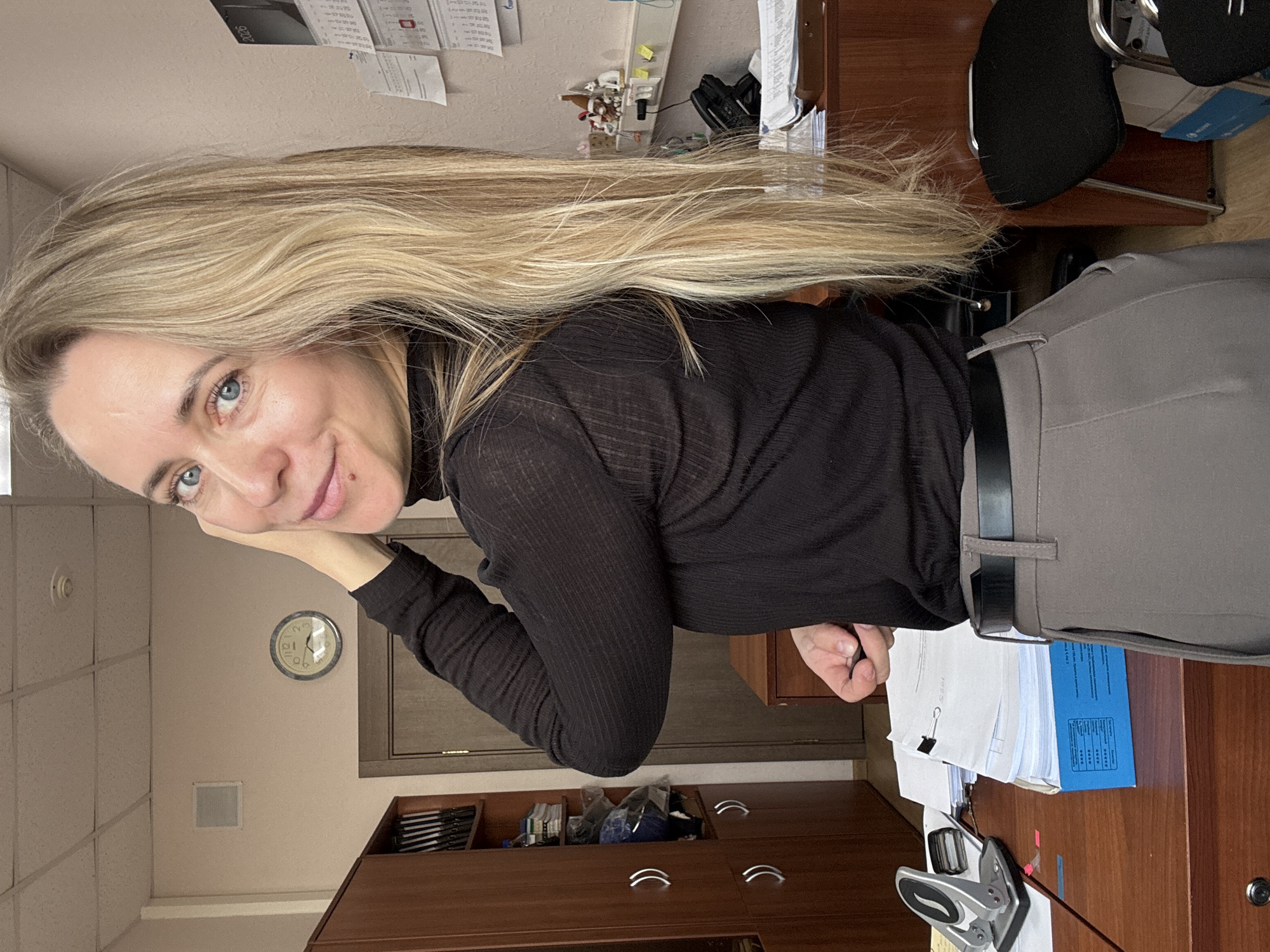 A fair-skinned woman with long, straight, blonde hair and blue eyes is captured in an office setting. She is seen from the chest up, leaning slightly on her right hand, and looking directly at the camera with a friendly, subtle smile. She wears a black, ribbed, long-sleeved turtleneck shirt paired with gray trousers and a black belt. Her hair cascades over her shoulders and back.
The background reveals an organized office environment, likely in Chaikovsky, Russia. To her left (viewer's right, considering the image's orientation), a dark wooden desk is cluttered with stacks of papers, blue folders, a silver hole puncher, and other office supplies. A black office chair with metal legs is partially visible behind her. Above the desk, a wall calendar shows "2026," and additional documents are taped to the wall. Further back, a round analog wall clock with black numbers and hands is visible, indicating a time close to twelve o'clock (the hour hand is near 12, minute hand near 10, suggesting 11:50 or 12:50). Below the clock, there are wooden storage cabinets or drawers. The ceiling features square acoustic tiles with visible grid lines. The overall scene suggests a routine day in an administrative or professional workspace.