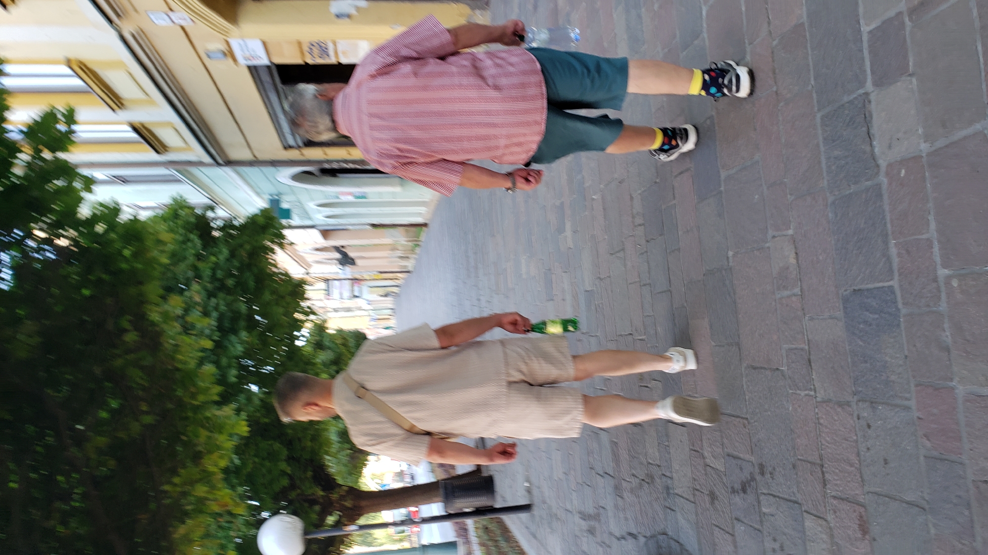 Two men are walking away from the camera on a cobblestone sidewalk in a European city, likely Prešov, Slovakia, based on the provided context. The man on the left wears a light-colored, short-sleeved collared shirt and matching shorts, with a light-colored strap across his shoulder. He holds a green glass bottle in his right hand. The man on the right wears a red and white striped, short-sleeved collared shirt and dark blue shorts. He has on patterned socks with athletic shoes and is carrying a clear plastic bottle of water in his right hand. They are walking down a street lined with buildings that have light-colored facades and dark-framed windows. Trees with green foliage are visible on the left side of the street, partially obscuring the view of the buildings. A lamp post with a white globe is also present on the left. The sunlight suggests it is daytime.