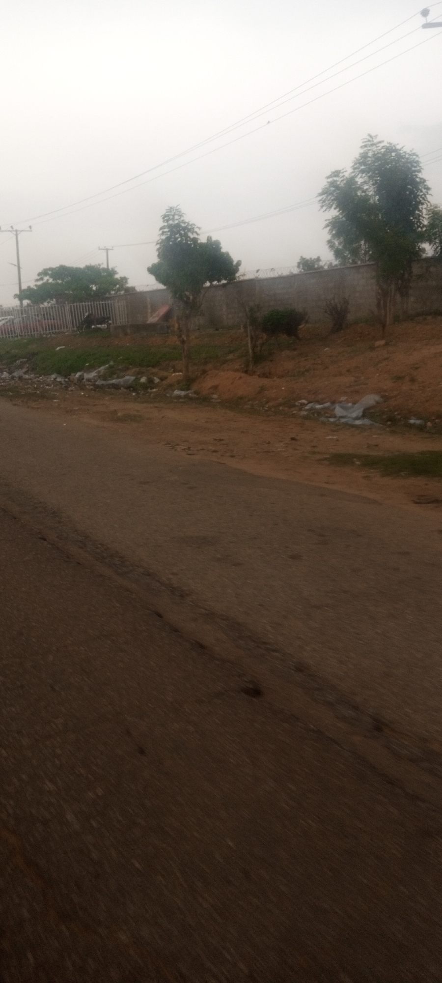The image depicts a street scene in Television, Nigeria, under an overcast sky. The focus is on the asphalt road in the foreground, which has visible wear and tear. To the side of the road, there's a grassy embankment with scattered trash and two young trees. A concrete wall with barbed wire runs along the top of the embankment. In the background, utility poles and more trees are visible, blending into the hazy atmosphere. There are no people visible in the image.