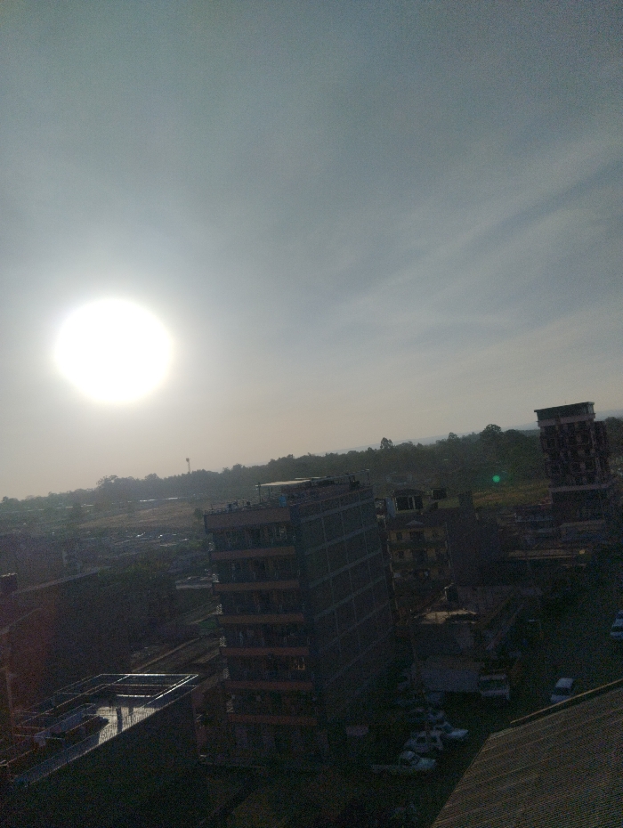 An elevated view captures a cityscape under a bright, hazy sun. Tall buildings dominate the scene, one with scaffolding-like exteriors. Shadows cast across the landscape hint at the early morning or late afternoon light. Vehicles are visible near the buildings. Distant trees and hills form a soft horizon, contrasting with the urban structures.
