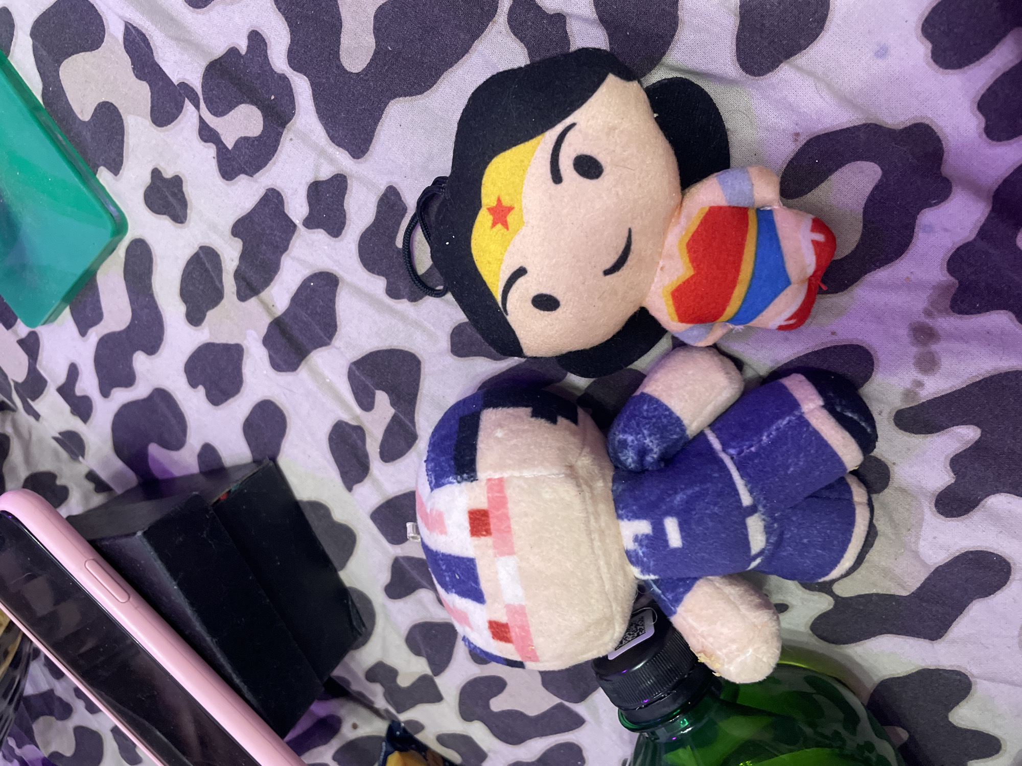 This image depicts two plush toys on a patterned surface, possibly a bedspread or fabric. One plush toy is Wonder Woman, recognizable by her costume including a red top with a gold trim, blue shorts, and a gold tiara with a red star. The other plush toy is a character with dark hair, a blue uniform with white accents, and a pixelated face with red and pink markings. To the left, a green bottle with a black cap is partially visible, along with a black box and a pink smartphone. The lighting in the image has a purplish hue, suggesting it might be artificial light or a specific setting.
The image does not contain any information or context that would allow for its location to be determined in Kokkorevo, Russia, or any other specific city. The scene is a still life arrangement of objects.