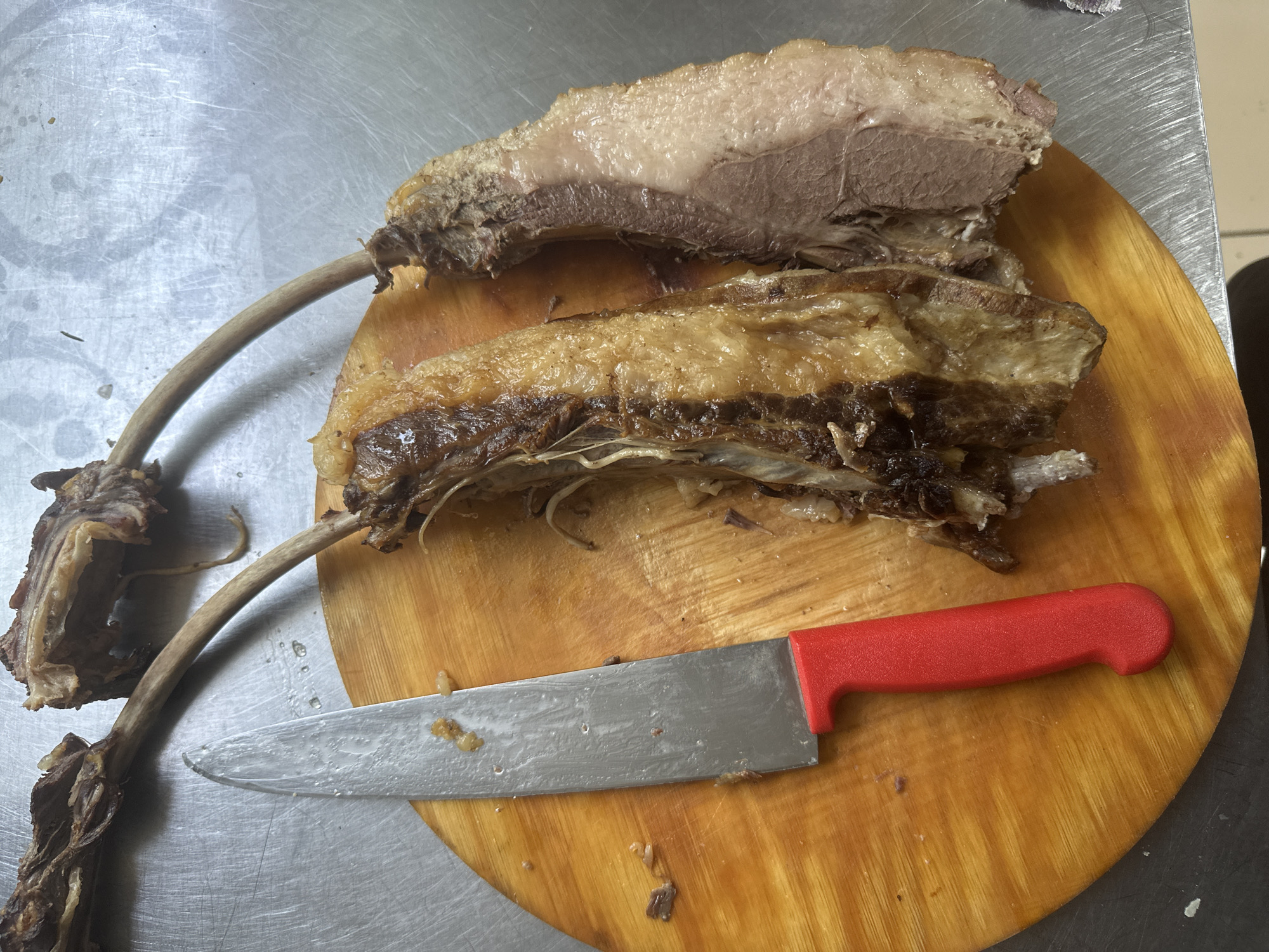 The image depicts two bone-in cuts of cooked meat on a round wooden cutting board. A large knife with a red handle lies to the left of the meat. The meat appears to be roasted or braised, with some browning and visible fat. The cutting board is placed on a metallic surface, likely a kitchen counter or table. There are no discernible elements in the image that would indicate a specific location within the city of Kostanay, Kazakhstan.