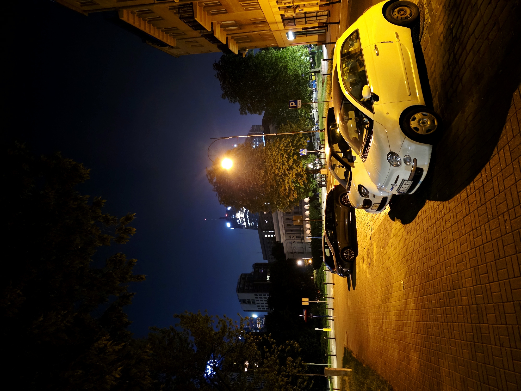 This is a nighttime exterior shot of a street in Warsaw, Poland, captured from a low angle. The scene features several cars parked along a sidewalk paved with interlocking bricks. The prominent subjects are two compact cars: a yellow Fiat 500 in the foreground on the right, and a white Fiat 500 parked behind it. A dark-colored car is also visible behind the white one.
The setting is an urban environment with buildings lining the street. One building, illuminated by artificial light, is on the left side of the frame, displaying rows of windows. In the background, the Warsaw skyline is visible, with tall buildings lit up against the dark night sky. Trees with dense foliage are also present, some casting shadows on the ground.
The time of day is clearly night, indicated by the darkness of the sky and the artificial lighting illuminating the scene. The weather appears to be clear, as there are no visible signs of rain or fog. The lighting from streetlamps and buildings creates strong shadows, adding depth to the image.
There is some visible text on the license plate of the white Fiat 500, which appears to be "WZ 214JH". There are also street signs visible, including a blue parking sign with a "P" symbol and Polish text, likely indicating parking regulations. Another sign, possibly a pedestrian crossing warning, is also present.
The overall mood is one of urban nighttime tranquility, with the illuminated cars and buildings creating a somewhat serene atmosphere despite the city setting. The low angle perspective emphasizes the cars and the architectural elements of the street.