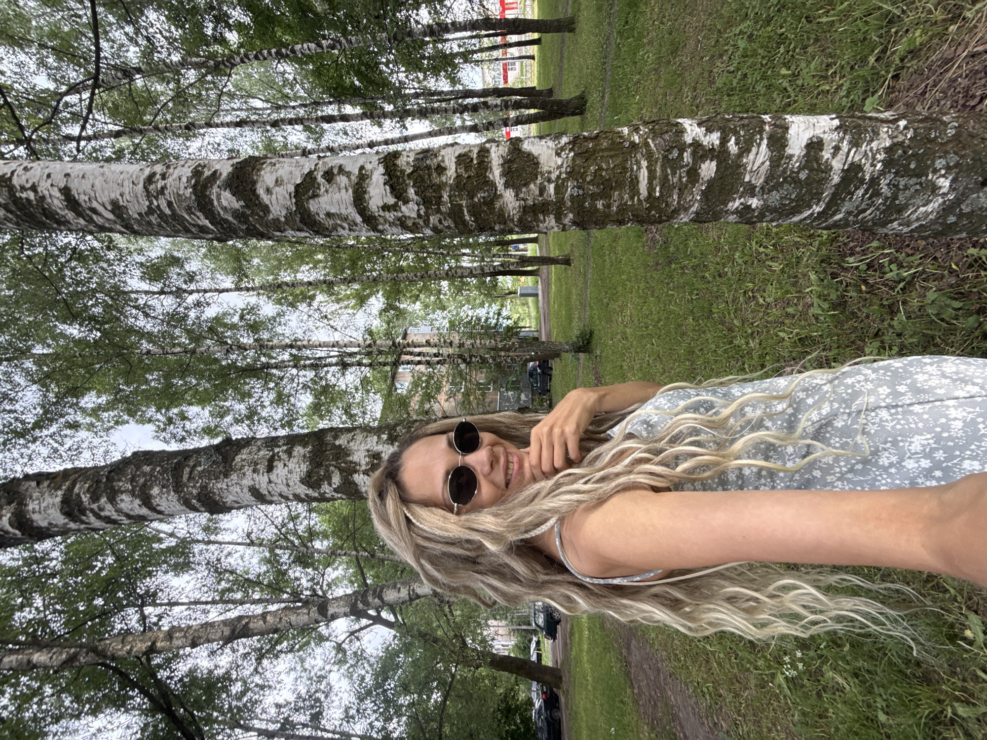 A woman with long, blonde, wavy hair and braids, wearing a light blue floral print dress and sunglasses, stands in a park-like setting. She is smiling and touching her chin with her right hand, holding a camera in her left hand. She is positioned in front of several birch trees with distinct white bark. The background shows a grassy area with more trees and some buildings and parked cars in the distance. The overall scene appears to be a casual outdoor photograph taken on a cloudy day.