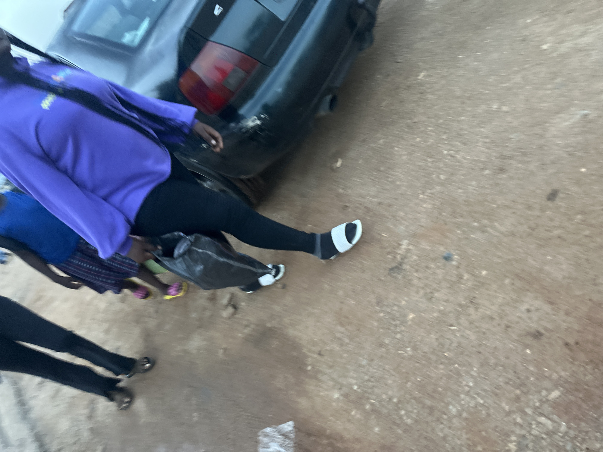 The media file shows a scene in Jos, Nigeria, focusing on a group of people standing near a black car. One person in the foreground, wearing a purple hoodie and black leggings, is visible from the torso down, with their feet in white and black sandals. They are holding a black plastic bag. Behind them, another individual is wearing a blue top and a striped skirt, with yellow and pink sandals. To the left, the legs of a third person in black leggings and sandals are visible. The car appears to be parked on a dirt or gravel surface, suggesting an outdoor setting. The lighting suggests it could be daytime, possibly late afternoon given the soft light. The overall impression is a candid, everyday moment. There is no visible text in the image.