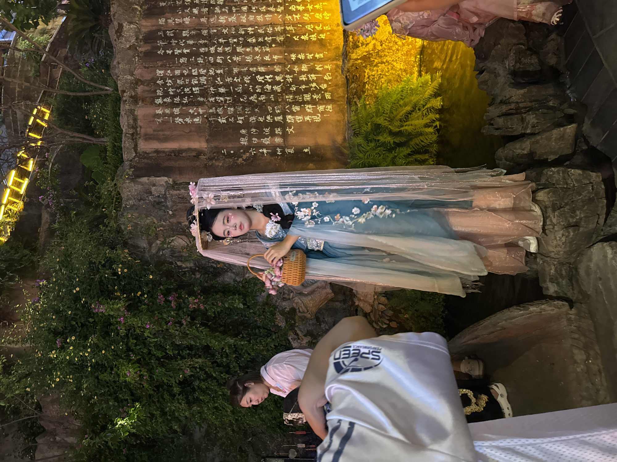 In Yuzhong District, China, a young woman dressed in a traditional blue and pink hanfu stands on rocks near a body of water. She wears a veil adorned with flowers and holds a woven basket filled with pink blossoms. Behind her, a stone wall is inscribed with Chinese characters, illuminated by yellow lights. To her left, another woman in a white shirt is partially visible, looking down. The environment includes lush green foliage and rocky terrain. The image captures a staged or cultural performance scene.