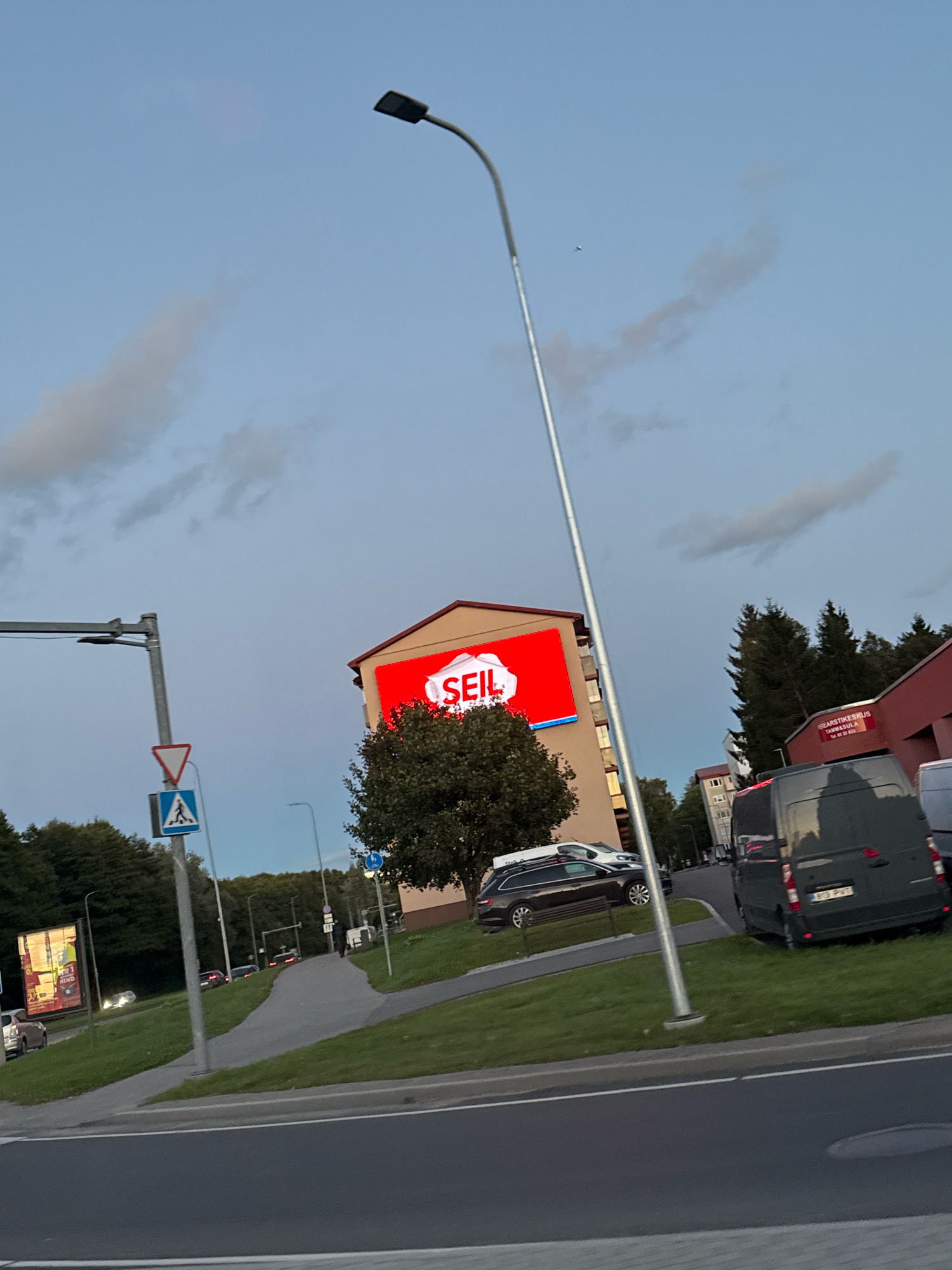 In Tallinn, Estonia, a street scene is captured at dusk. A tall, grey lamppost dominates the foreground, its light fixture angled towards the viewer. To the left, a traffic light pole with multiple signals is visible. A street sign with a pedestrian crossing symbol and a triangular warning sign is mounted on a pole. In the middle ground, a multi-story building displays a large red billboard with the word "SEIL" in white lettering. Adjacent to the building, a tree provides shade. Several cars are parked along the street, including a dark colored station wagon and a dark grey van with a license plate reading "873 PVT". In the background, more trees and buildings can be seen, along with other vehicles and streetlights. The sky is overcast with a hint of blue.