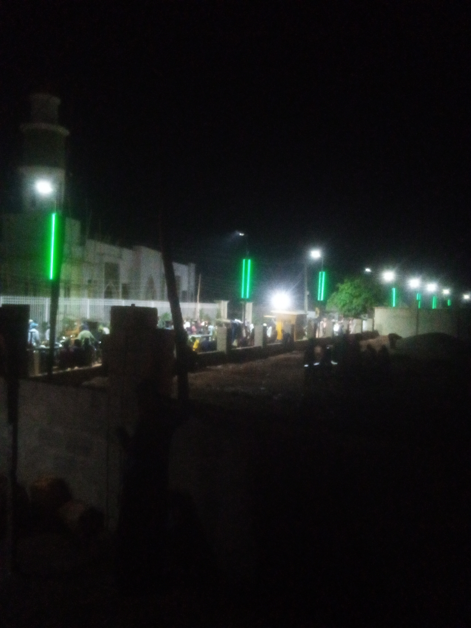 In Tundun Wada West, Nigeria, at night, a mosque's minaret is visible on the left, illuminated by a white light at its top and a bright green light further down. The mosque structure itself is light-colored and rectangular, with windows. In front of the mosque, a white fence encloses an area where many people are gathered, appearing to be engaged in evening prayers or a religious gathering. They are seated or prostrating on the ground, with prayer mats visible. Several vertical green lights are positioned along the roadside, illuminating the scene. The foreground is dark, with some shadowy figures and structures. The overall impression is a nighttime religious event taking place outside a mosque in Nigeria.