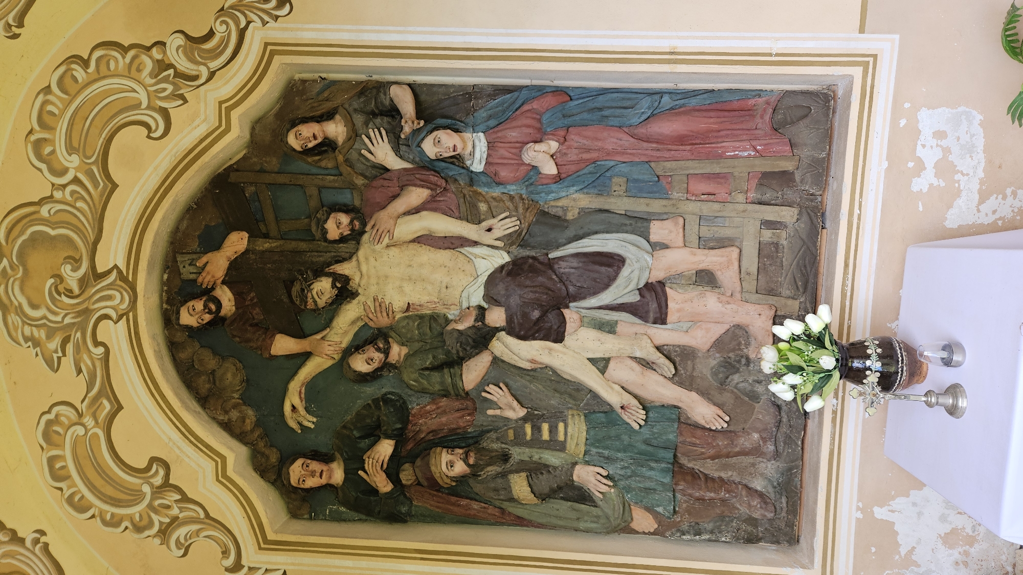 The image depicts a religious relief carving, likely within a church setting, possibly in Prešov, Slovakia. The carving shows the Deposition of Christ, with multiple figures surrounding the body of Jesus. A woman in a red robe and blue mantle lies above Christ's body, possibly representing the Virgin Mary. Several men support and cradle the body. The scene is framed by an ornate, yellow border, possibly stucco. To the right of the carving, a table covered with a white cloth holds a vase of white flowers and a silver crucifix. The wall adjacent to the carving is light pink and shows signs of age.