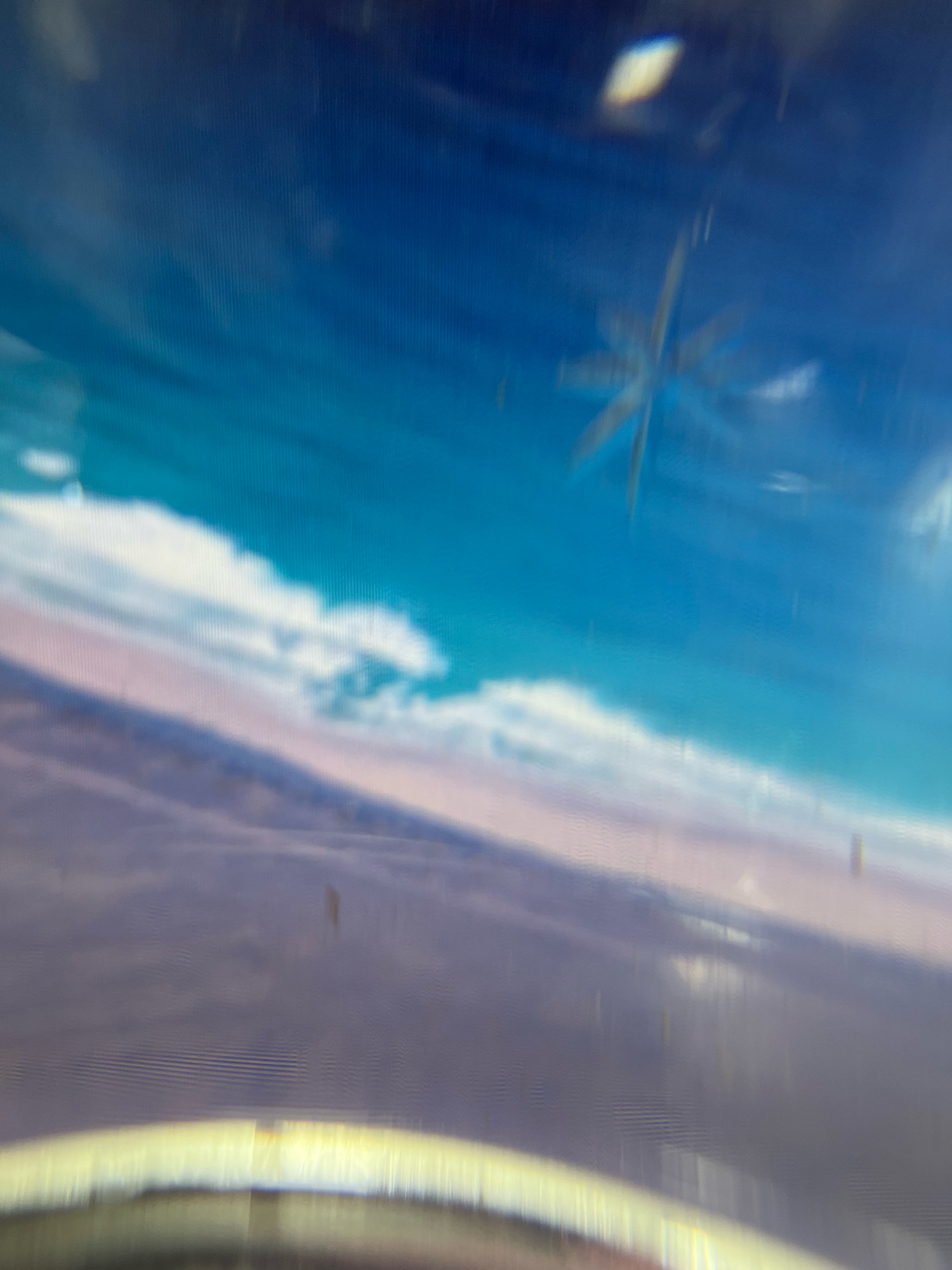 The image displays a digital screen showing a blurry, abstract scene. The upper portion of the screen is a gradient of deep blue to turquoise, suggesting a sky. There are faint white shapes resembling clouds. In the middle right of the screen, a blurred, star-like or snowflake-like shape is visible, possibly a reflection or a digital artifact. The lower portion of the screen transitions to muted purples and grays, depicting what could be a landscape or further cloud formations. A bright, horizontal band of yellow light is present at the very bottom, with a distorted, panoramic effect, suggesting the edge of the screen or a reflective surface. The image appears to be taken of a screen, as indicated by a slightly curved, reflective yellow border at the bottom and a faint grid pattern visible across the surface. There are also small, irregular dark spots scattered across the screen, likely dust or smudges. The scene has a dreamlike or out-of-focus quality. No specific landmarks or context related to Manesar, India, are discernible within the image itself.