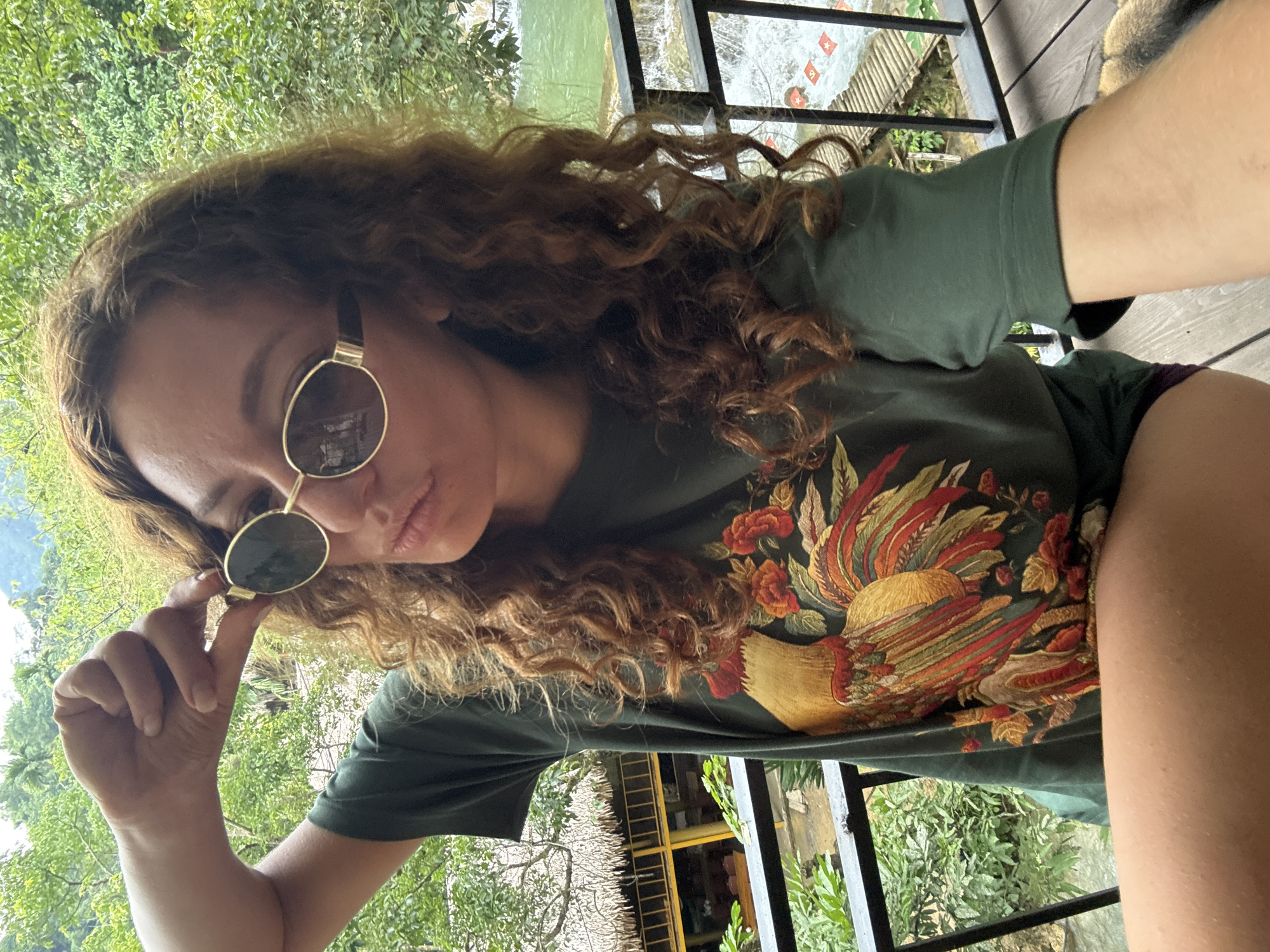A young woman with curly brown hair and sunglasses is taking a selfie on a wooden balcony. She is wearing a dark green t-shirt with a large embroidered rooster and floral pattern. Her right hand is raised, adjusting her sunglasses. In the background, lush green trees and foliage are visible, with a glimpse of a thatched-roof building and a waterfall with a wooden walkway. The scene suggests a tropical or mountainous environment, consistent with locations like Cổ Lũng Commune, Vietnam.