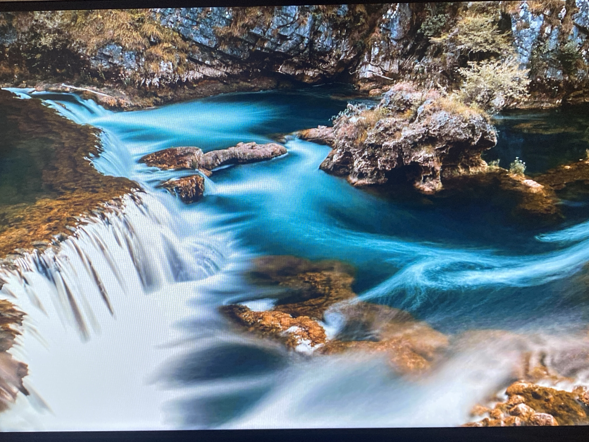 The media file presents a stunning natural landscape, captured with a long exposure photographic technique, featuring a waterfall and a vibrant river system. No people or visible text are present in the image.
The scene is dominated by the dynamic movement of water. On the left side, a series of cascades or a multi-tiered waterfall flows over dark, rugged rock formations, rendered into a silky, blurred white by the long exposure. This water then feeds into a wide, turquoise-blue pool that snakes through the center of the frame. The water in the pool exhibits various shades of blue and green, with swirling patterns created by the currents, also softened by the photographic technique.
Several large, weathered rock formations, covered with earthy-toned moss and sparse vegetation, punctuate the water's flow. One prominent rock island, situated on the right side, features small green plants and appears to be undercut by the water. The background consists of tall, rugged cliffs and rocky banks, adorned with patches of brown and green foliage, suggesting a dense, natural environment.
The notable details include the striking contrast between the smooth, ethereal water and the sharp, textured rocks and vegetation. The overall color palette is rich, with vibrant blues and greens from the water contrasting with the warm browns, oranges, and deep grays of the rocks and earth. The lighting suggests natural daylight, possibly indirect or overcast, which enhances the colors and the artistic effect of the long exposure. The atmosphere is one of serene natural beauty and the continuous, powerful flow of nature. The image depicts a wild, untamed riverine environment, and while an external context of Saint Petersburg, Russia, is provided, the visual elements within the image do not offer specific geographic cues for that urban location.