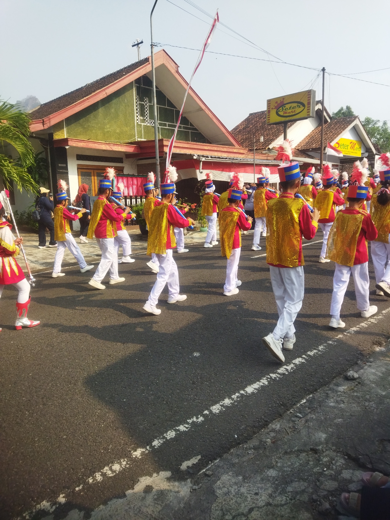 In the city of Wonogiri, Indonesia, a marching band of children, dressed in vibrant red and gold uniforms with blue and white plumed hats, parades down a street. The children are playing instruments, possibly flutes or clarinets, and are marching in formation. The street is paved with asphalt and marked with a white line. On the left side of the street, there are houses with red-tiled roofs and green-painted walls. A sign for "Seter Mart" is visible on the right side of the street. The sky is clear and the sun is shining, casting shadows on the ground. The overall scene suggests a festive or celebratory event.
