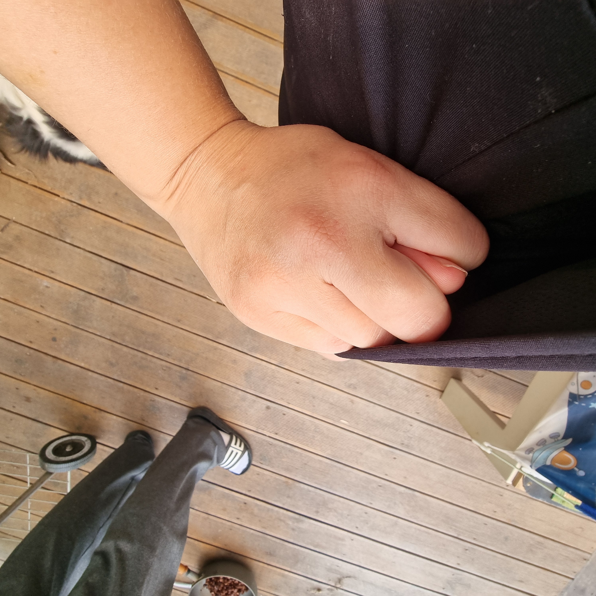 The image depicts a close-up view of a person's hand holding black fabric, likely clothing, against a backdrop of a wooden deck. The hand is in focus, with visible skin texture. Partially visible in the lower right portion of the frame are the legs and feet of the same person, wearing dark pants and slide-on sandals. A small wheeled cart is positioned near the feet. In the far right background, a metal pot containing small, dark objects, possibly food, is visible. The wooden deck appears to be outdoors, with parallel planks running diagonally across the frame. No specific location within Dunnae-myeon, South Korea, can be identified from the image.