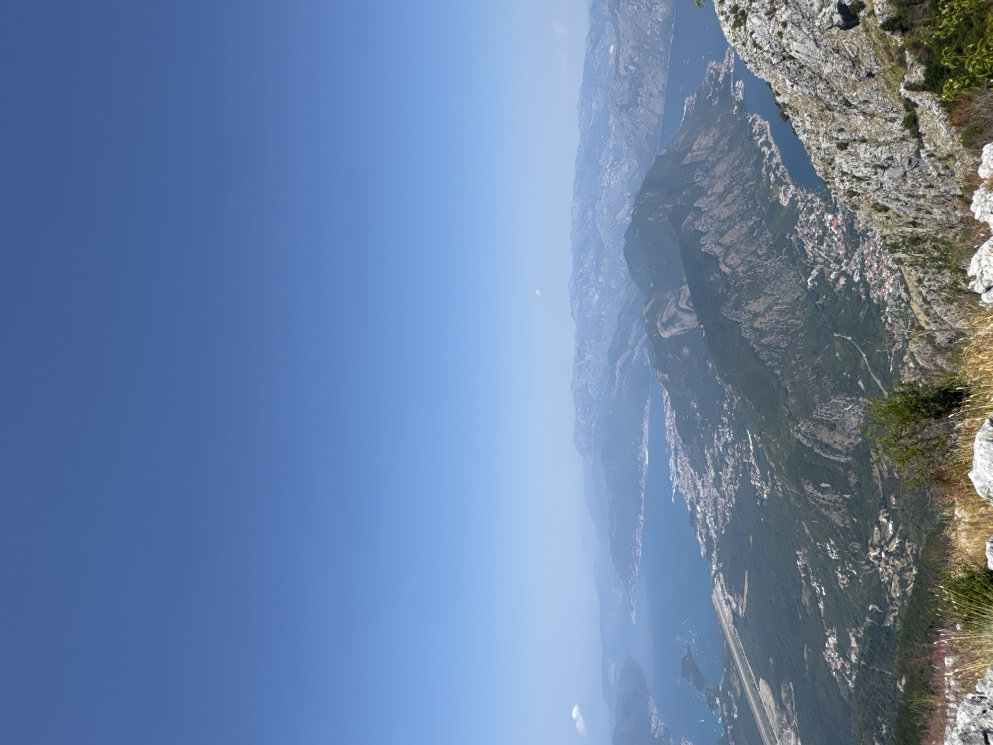 This image provides a high-angle panoramic view of a bay and surrounding mountainous terrain, likely captured from a summit. The sky is a clear, pale blue. In the mid-ground, a body of water, appearing to be a bay, is visible with a distinct shoreline featuring scattered settlements and a linear feature that resembles an airport runway. Dense green vegetation covers the lower slopes of the mountains. The foreground consists of rugged, rocky terrain with sparse dry grasses and a small green shrub. The distant background is dominated by layered mountain ranges fading into a hazy, lighter blue. The scene suggests a clear, sunny day with good visibility, albeit with atmospheric haze softening the distant views. This landscape is consistent with the geography of the area around the Bay of Kotor in Montenegro, and the specific viewpoint could be from a mountain overlooking the town of Kotor or Tivat, in the vicinity of Kuk.