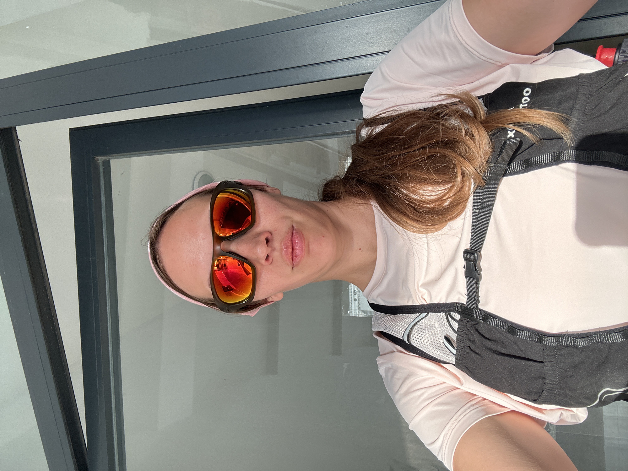 A woman wearing a pink headband, mirrored sunglasses, a light pink long-sleeved shirt, and a black running vest poses for a selfie. She stands near a gray-framed glass door or window. The reflection in the sunglasses suggests an outdoor scene in the daylight. The location may be Fethiye, Turkey.