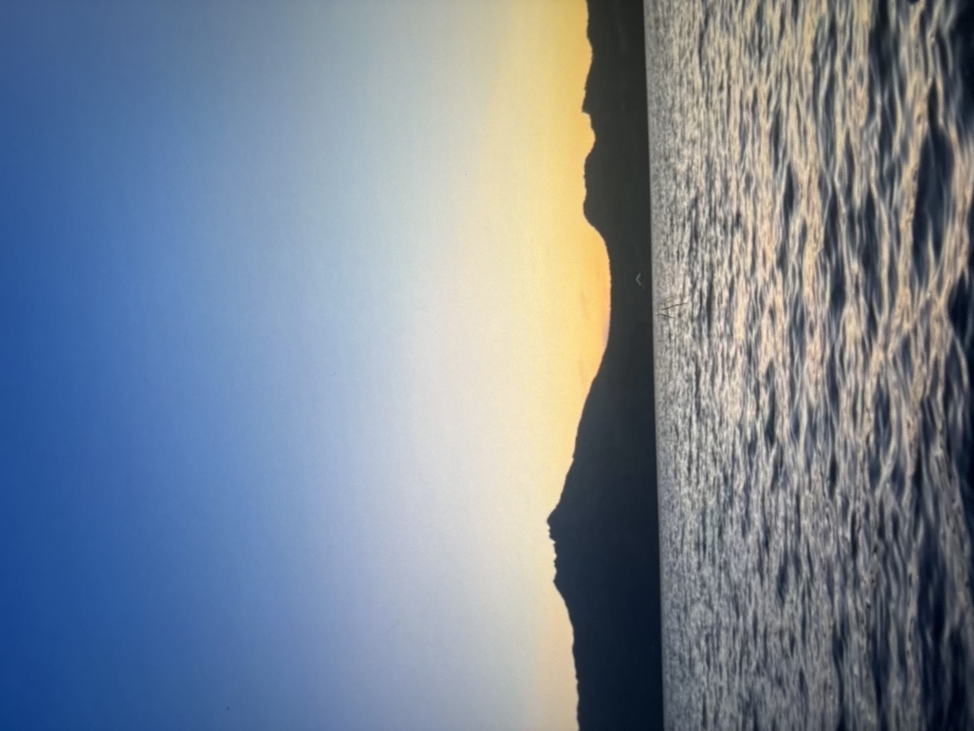 The image depicts a serene lake or body of water in Omsk, Russia, at either dawn or dusk. The sky transitions from a deep blue at the top to a warm yellow and orange gradient near the horizon, indicating the presence of the sun just above or below the visible line. Silhouetted against this colorful sky are several mountain ranges or large hills, their dark outlines sharply defined against the softer hues of the sunset. The water in the foreground is calm, with subtle ripples reflecting the colors of the sky, creating a peaceful and tranquil atmosphere. The scene is devoid of any human presence or man-made structures, emphasizing the natural beauty of the landscape.