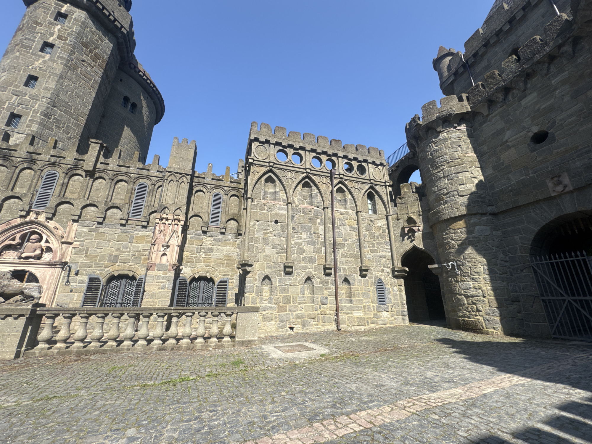 The image captures the exterior of **Löwenburg Castle**, a medieval-style castle located in **Kassel, Germany**. The castle is constructed of stone and features imposing towers, battlements, and arched windows with shutters. The foreground shows a cobblestone courtyard with a patterned design, and the sky is a clear, bright blue. The architecture is detailed and appears well-maintained. There are no people visible in the image, and the scene is static, focusing solely on the architectural elements of the castle and its surroundings.