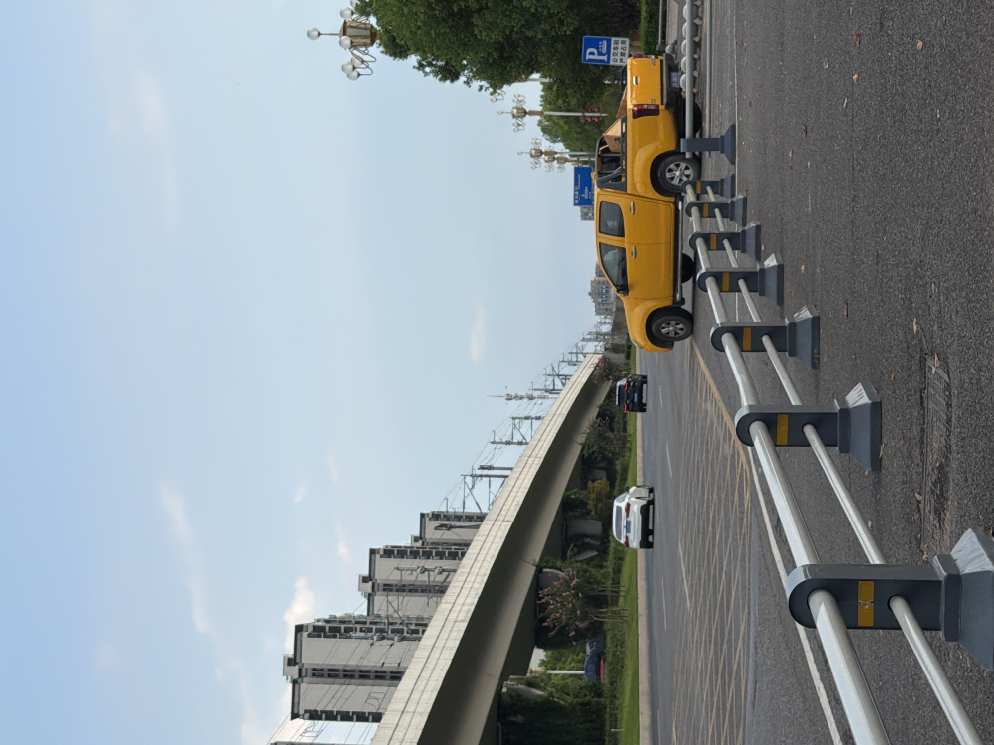 The image captures a street scene in Jiangjin District, Chongqing, China, under a clear sky with scattered clouds. In the foreground, a row of grey and silver traffic barriers separates the viewer from the road. On the road, a bright yellow pickup truck is parked, partially obstructing a blue traffic sign that indicates "Public Transportation Station, No Parking." Further down the road, a white sedan and a dark-colored SUV are in motion. An elevated structure, likely an overpass or a train track, stretches across the mid-ground, supported by concrete pillars. Tall, multi-story residential buildings with visible windows line the left side of the elevated structure. The right side of the image features lush green trees and ornate streetlights. The overall environment suggests an urban setting, possibly on the outskirts or a developing area of Jiangjin. No people are visible in the image.