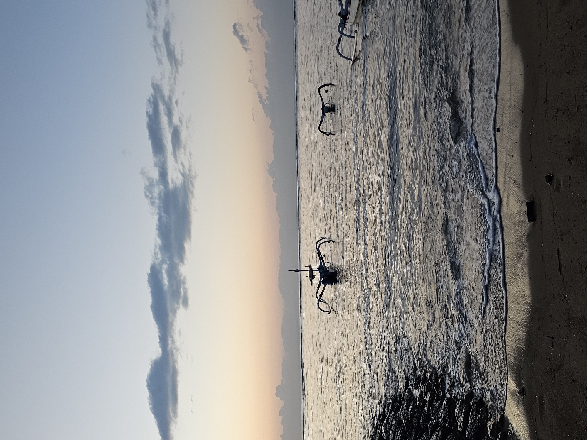 The image depicts a serene beach scene at sunrise in Denpasar, Indonesia. The calm ocean water laps gently onto a sandy shore, which is bordered by a rocky embankment on the left side of the frame. Three traditional outrigger fishing boats, known as jukungs, are visible in the water. One jukung is positioned in the foreground, slightly to the left of center, with its outriggers extended. Another jukung is further out in the water to the right, and a third is visible in the far right of the frame. The sky is a soft gradient of pale blue and peach, with wispy clouds scattered across the horizon. The overall atmosphere is peaceful and tranquil, characteristic of a sunrise over the ocean. There are no people visible in the image.