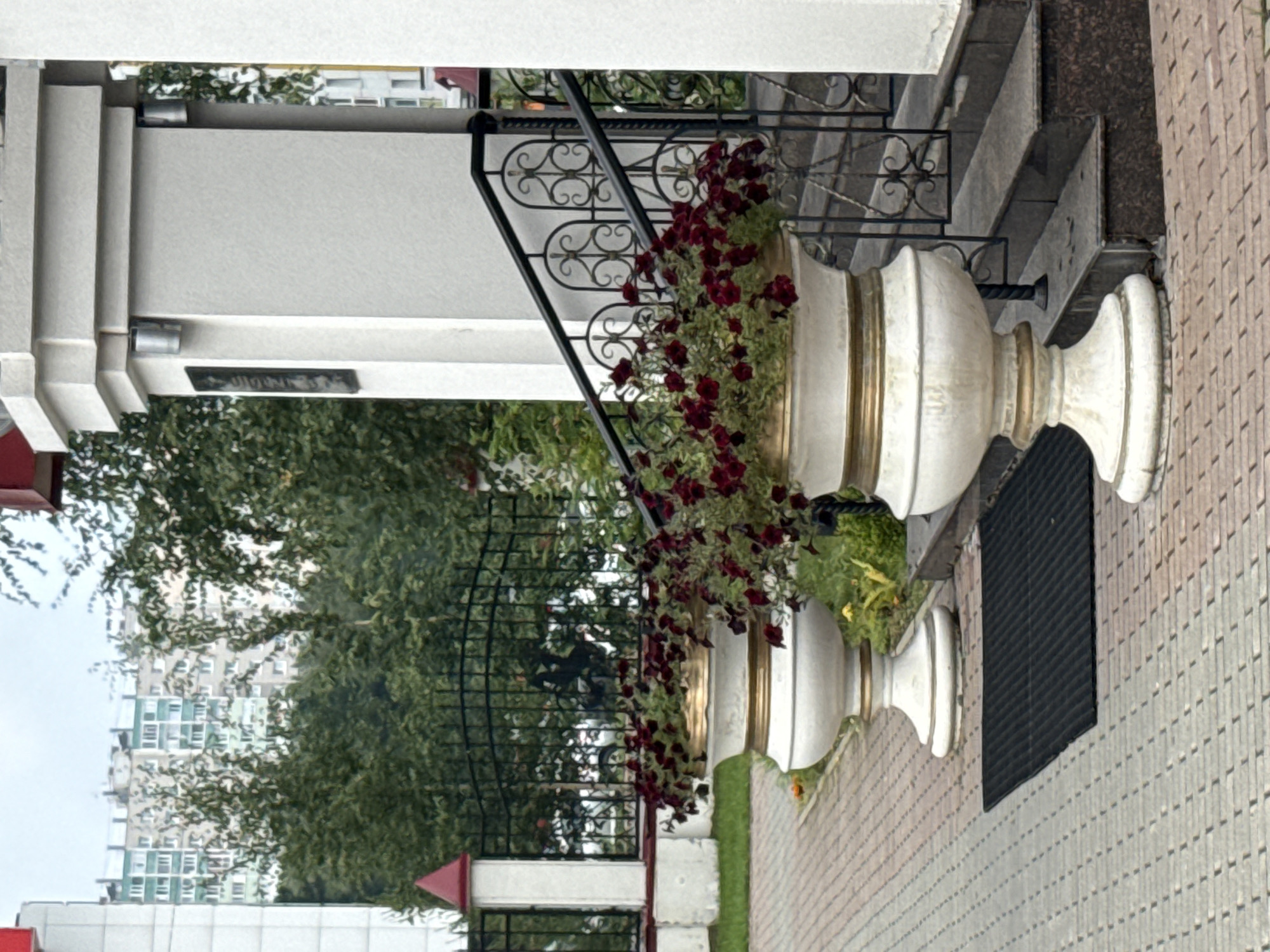 An eye-level outdoor medium shot captures a paved walkway in front of a building with a stone pillar and steps. In the foreground, two large, ornate white planters filled with dark red flowers flank a black doormat. The planters have gold banding. The walkway is made of tan and gray interlocking bricks arranged in a diagonal pattern. A black metal fence with decorative scrollwork runs behind the planters, separating the walkway from a grassy area and trees. Beyond the trees, a tall, multi-story apartment building with a light green and white facade is visible under a cloudy sky. The building features numerous windows and a flat roof. To the right of the image, a set of stone steps leads up to an entrance, with a black metal railing. A white, textured wall and a large stone pillar are also present on the right side of the frame. The overall impression is one of an urban setting with decorative landscaping.