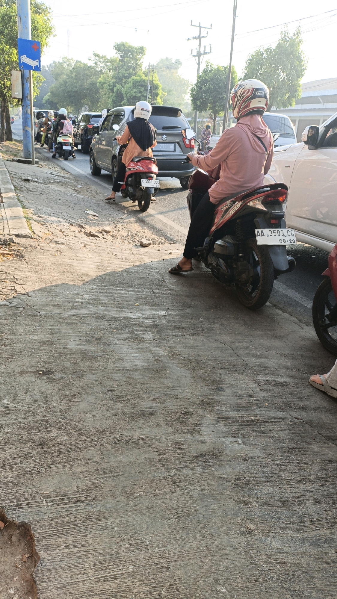 In Kuwasen, Indonesia, at midday, a traffic jam is captured on an asphalt road. The scene is dominated by several motorbikes and cars, creating a bottleneck on the road. A blue sign with a white hospital symbol indicates the proximity of a medical facility. On the right side of the image, a woman wearing a pink hooded jacket and a white helmet is riding a red motorbike. She is looking forward, and her feet are resting on the footpegs. Her motorbike displays a license plate with the characters "AA 3503 CG" and "08-29". Another motorbike with a rider wearing a white helmet and a black headscarf is in front of her, also facing forward. The road is lined with trees and buildings visible in the background, suggesting an urban or suburban environment. The ground in the foreground is a concrete surface with a textured pattern, showing signs of wear and tear. The lighting indicates a bright, sunny day.