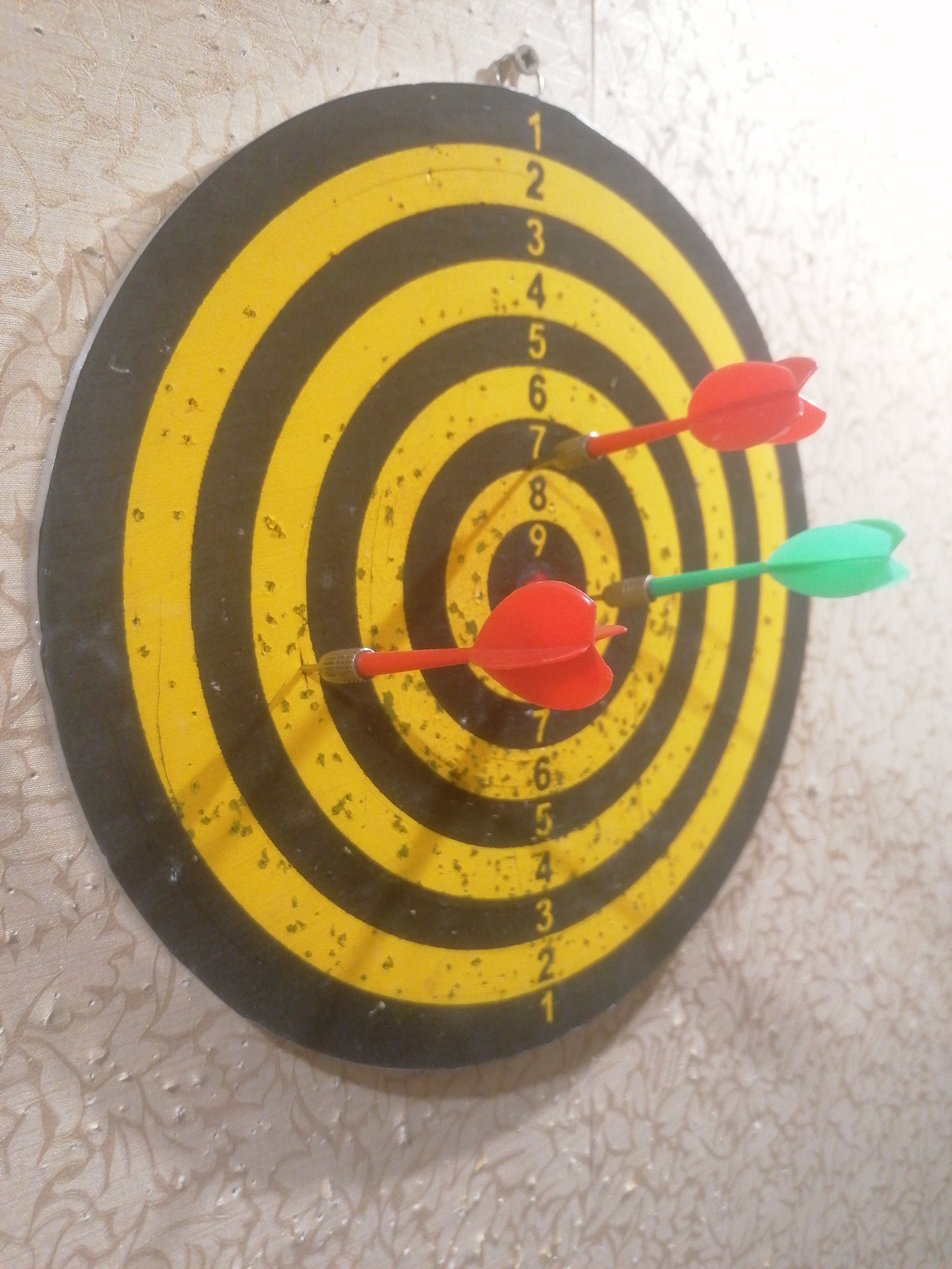 In Angarsk, Russia, a dartboard is mounted on a patterned wallpapered wall. The dartboard is circular with alternating black and yellow rings. Three darts are stuck in the board. One red dart is in the outer black ring, a second red dart is in a yellow ring, and a green dart is in another yellow ring. The darts are positioned such that they create a visual line, with the red dart in the upper yellow ring being the highest, followed by the green dart in the lower yellow ring, and the red dart in the outer black ring being the lowest. The lighting in the scene appears to be coming from the right, creating a bright highlight on the upper right portion of the image.